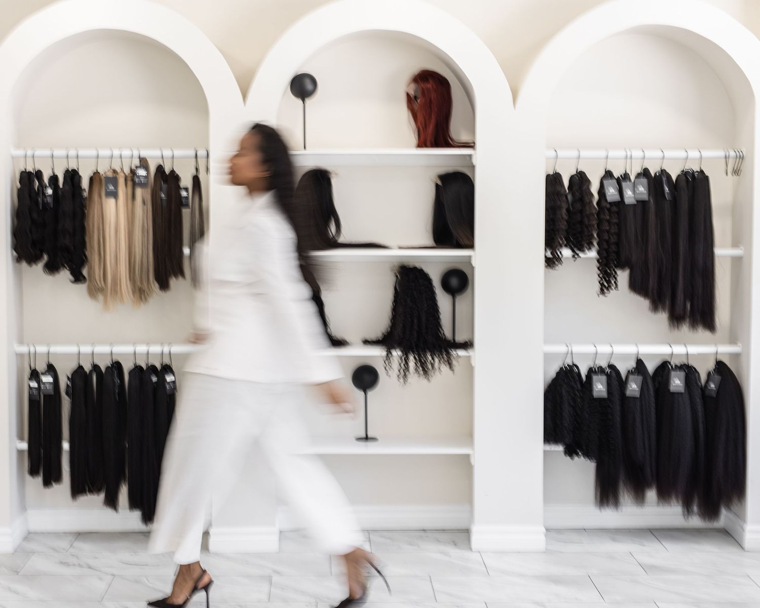 Woman in white outfit walking past arched shelving displaying black garments and hair pieces in a boutique.