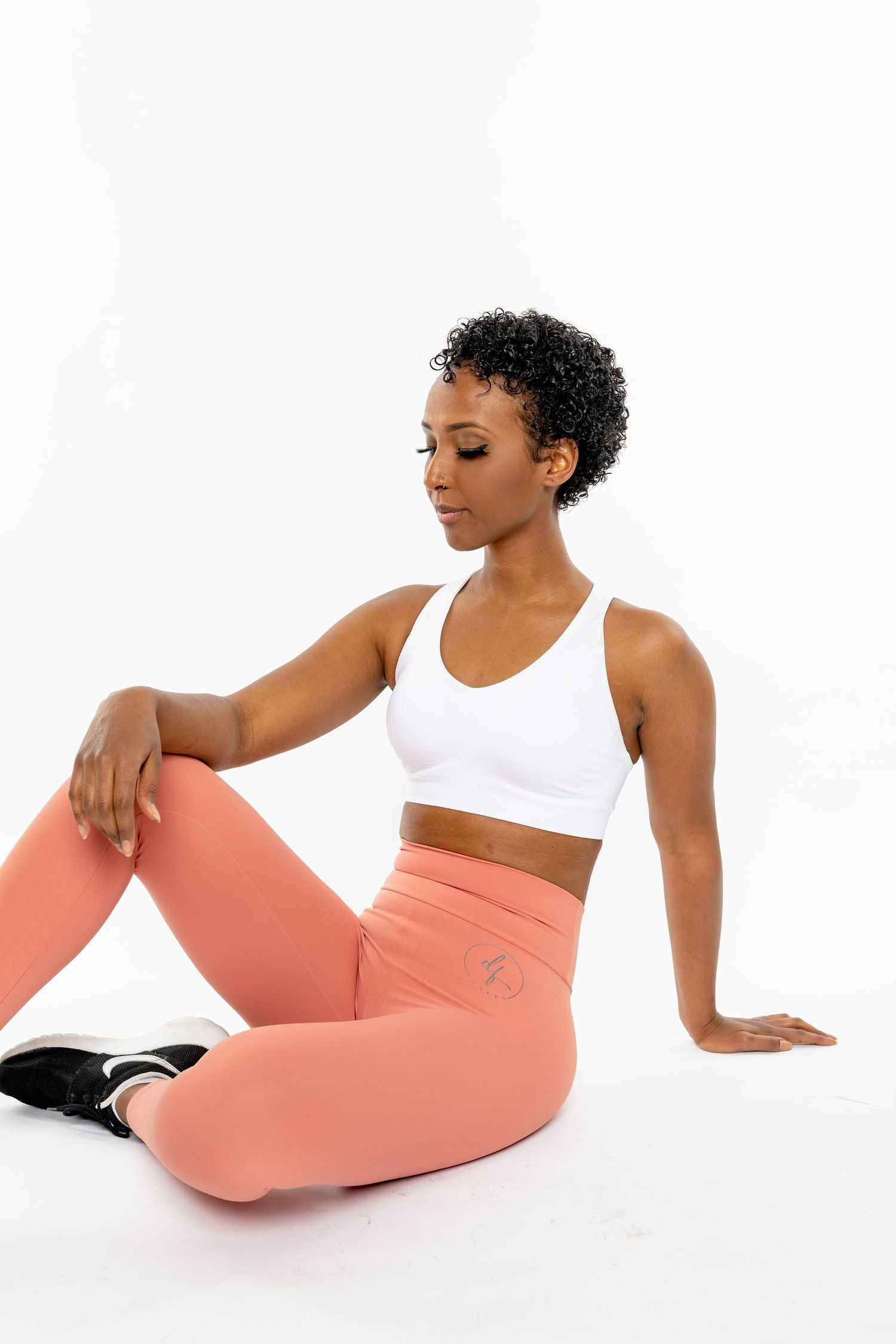 Woman in white sports bra and pink leggings sitting on white floor in athletic wear.