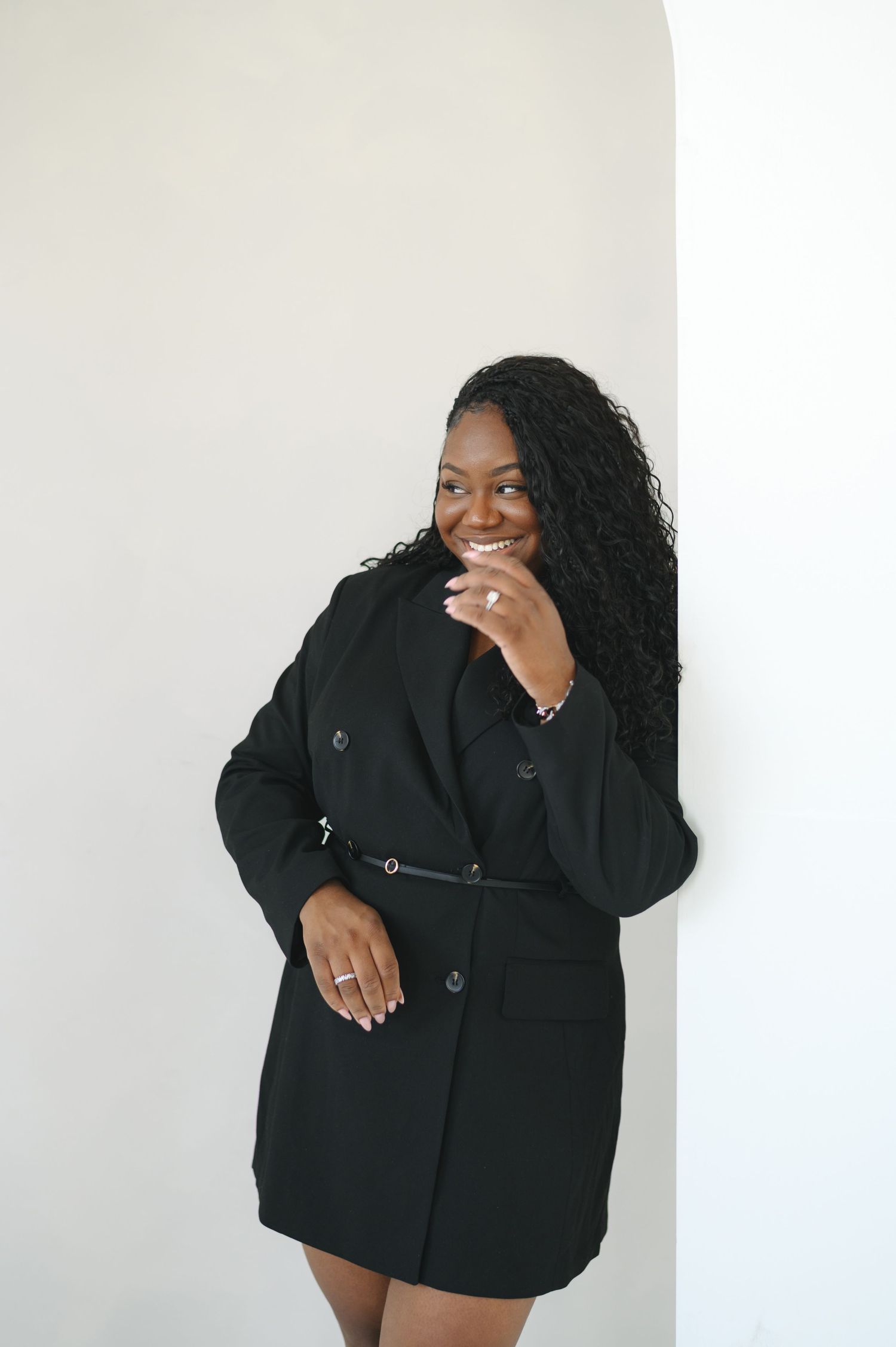 Model in black blazer dress poses playfully against white backdrop during studio photoshoot.