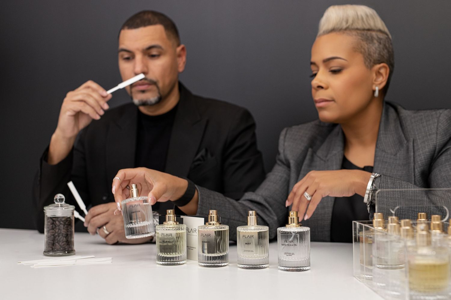 Two perfumers smelling fragrance strips beside glass candle jars on a white table in a studio setting.
