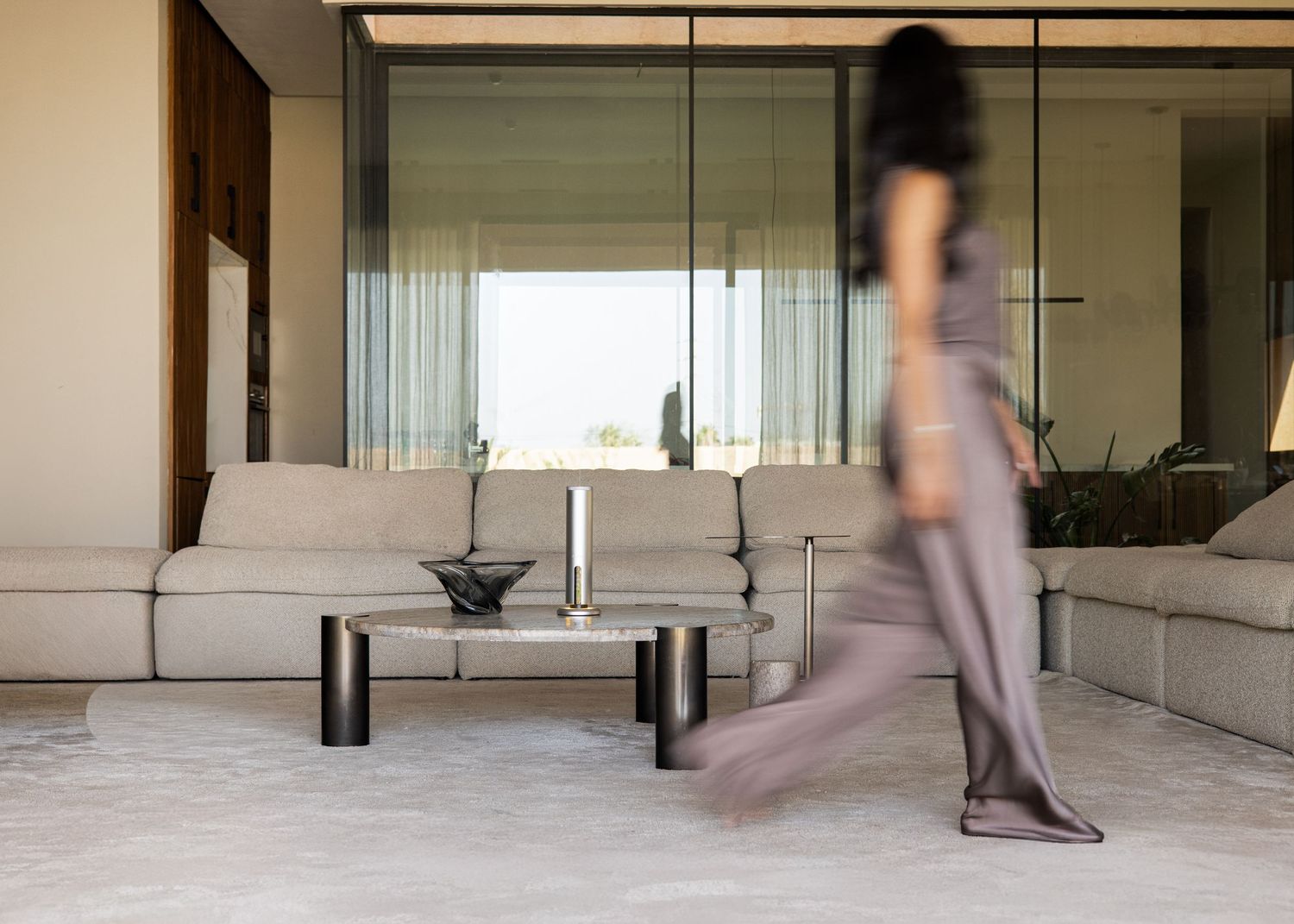 A blurred figure walks through a modern living room with a beige sofa, coffee table, and large glass windows.