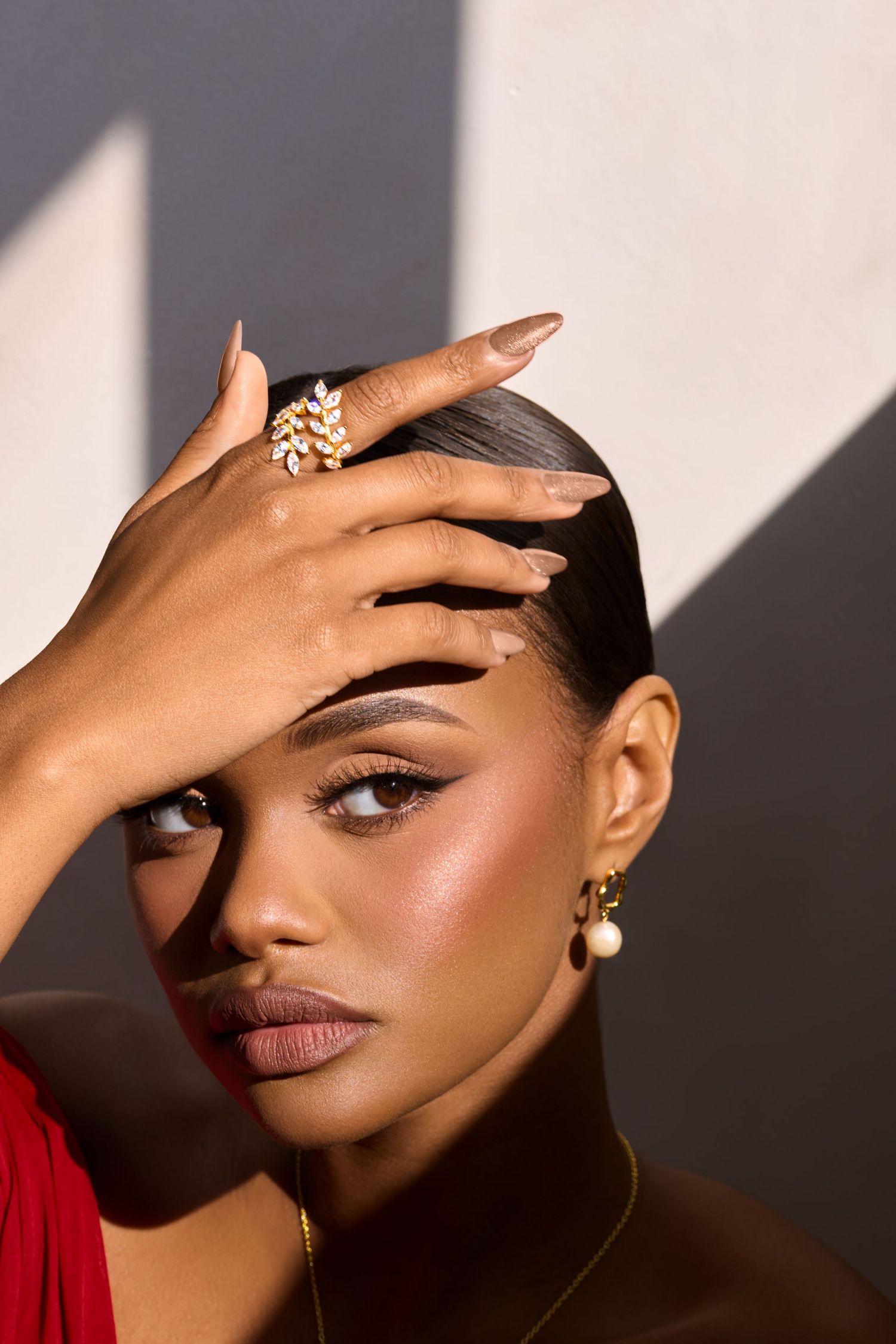 A woman with glowing skin poses with a jeweled ring and pearl earrings, hand raised to forehead in dramatic light.