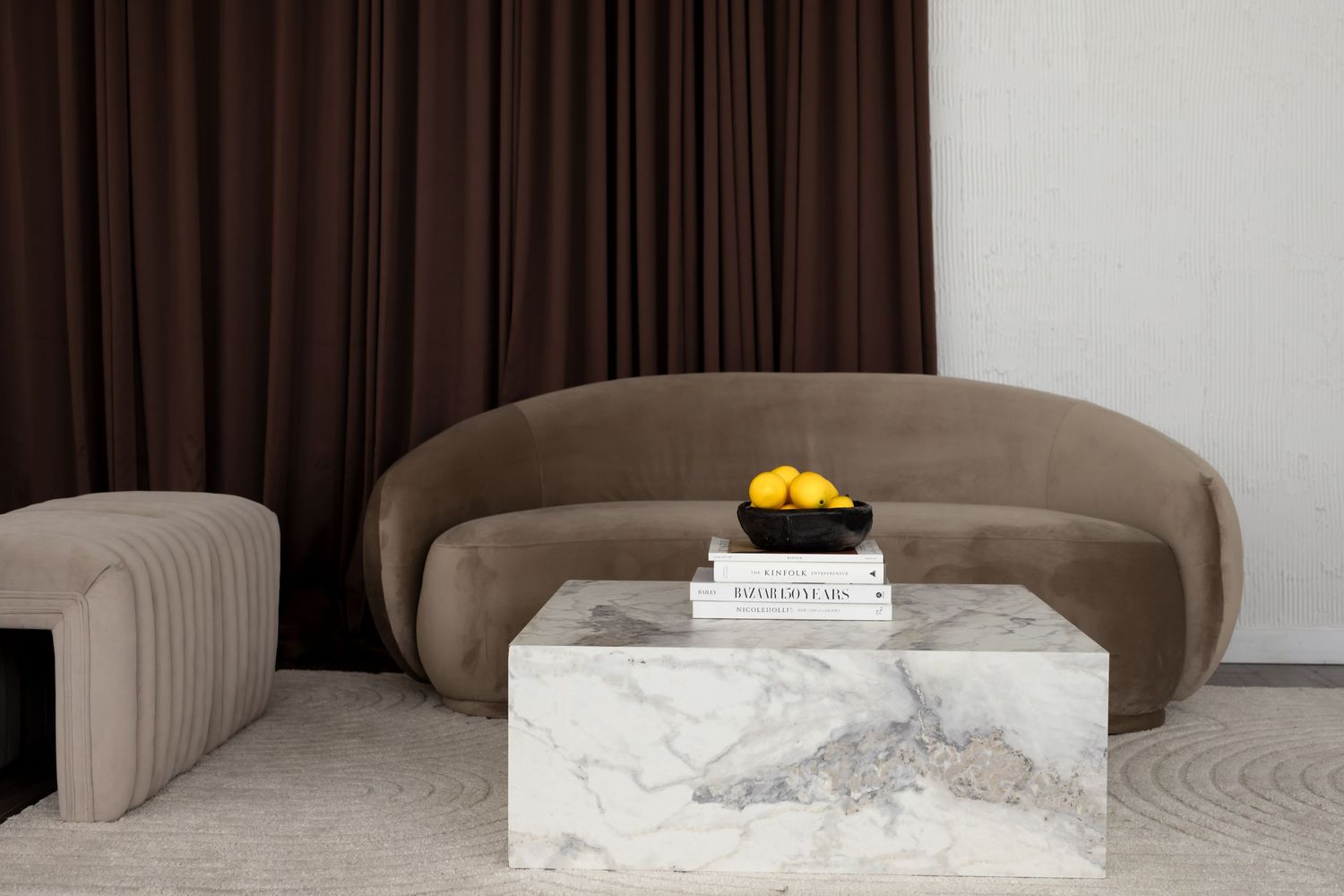 Curved brown velvet sofa with marble coffee table, black bowl of lemons, and books in a minimalist living room.