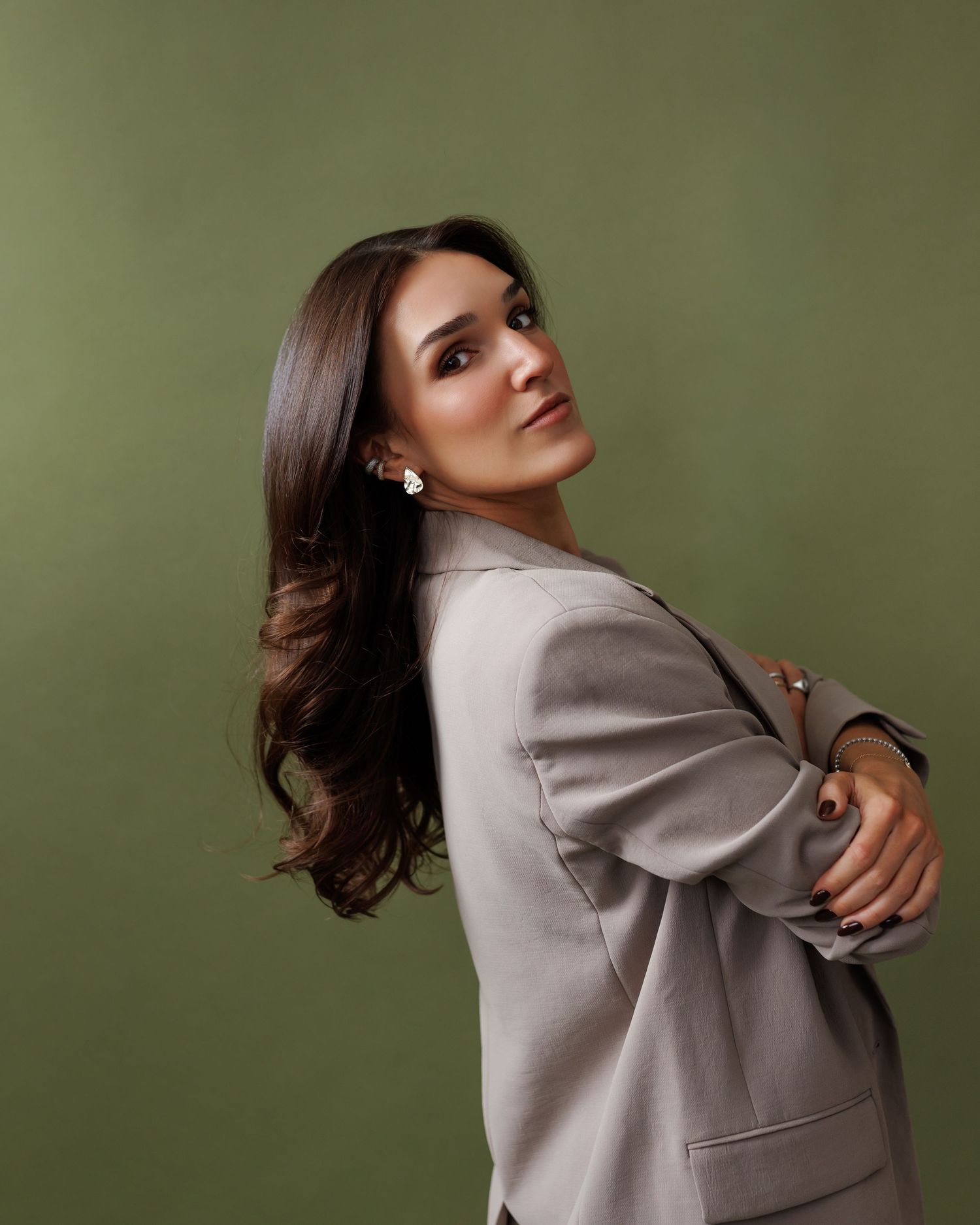 Woman in gray blazer poses confidently against green background with arms crossed, long dark hair, pearl earrings.