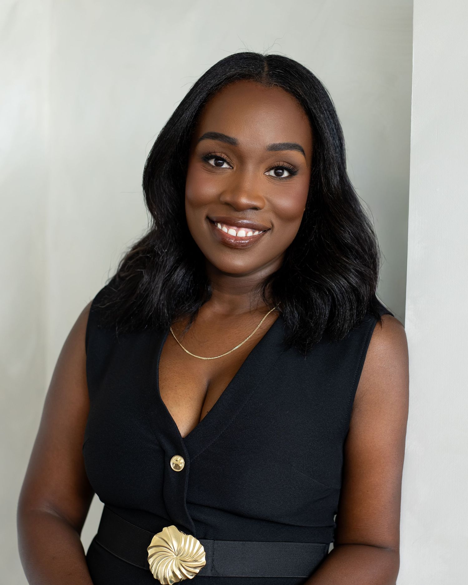 Professional headshot of a smiling young Black woman in a black sleeveless top with gold accessories.