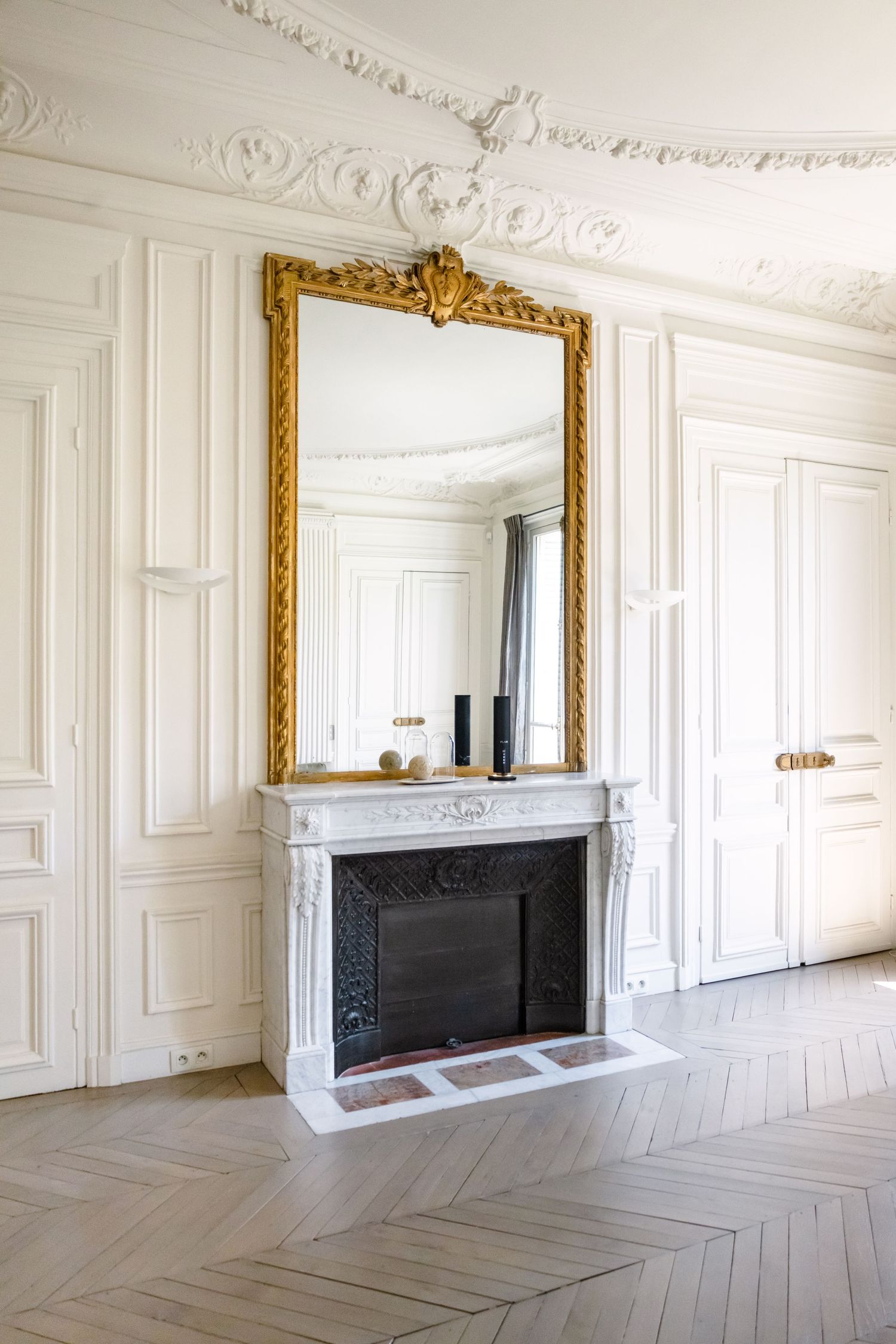 Elegant French interior with white marble fireplace, ornate gold mirror, herringbone parquet floor, and decorative crown molding.