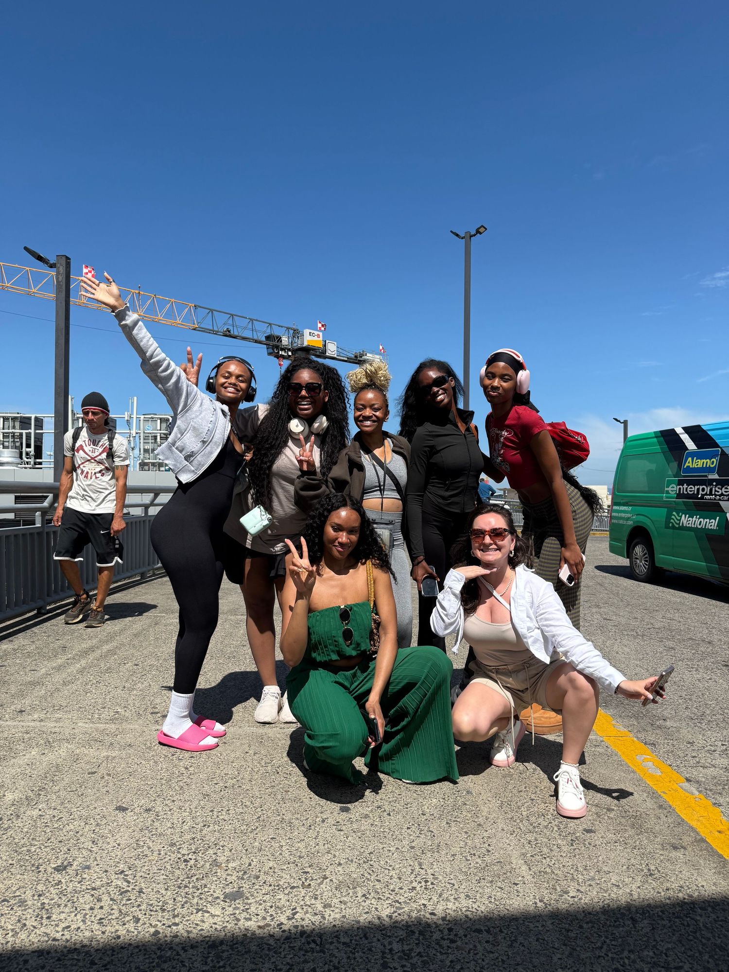 A group of friends pose playfully in front of a green barrier at an outdoor location on a sunny day.