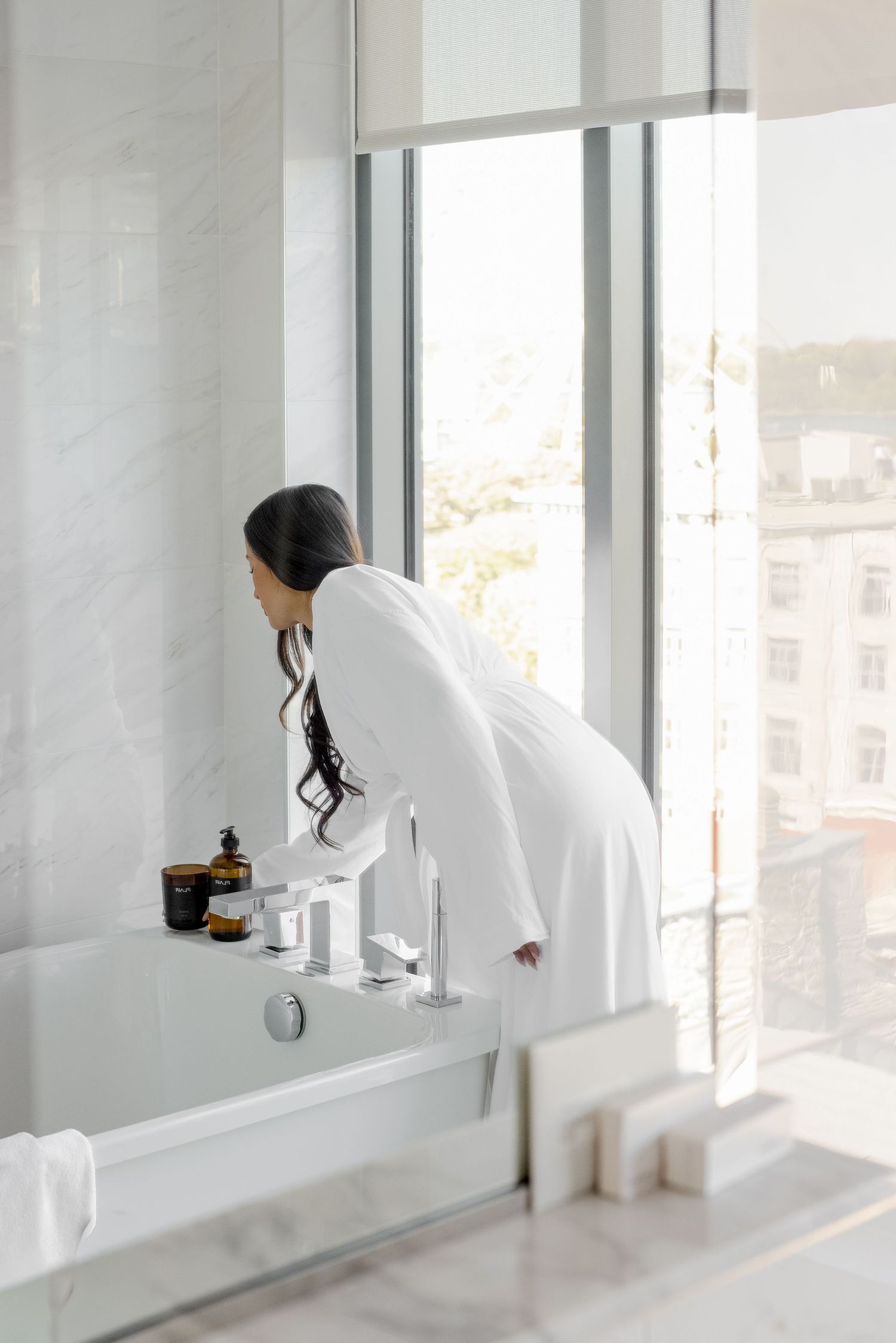 Woman in white robe leaning over a luxury bathtub in a marble bathroom with large windows.