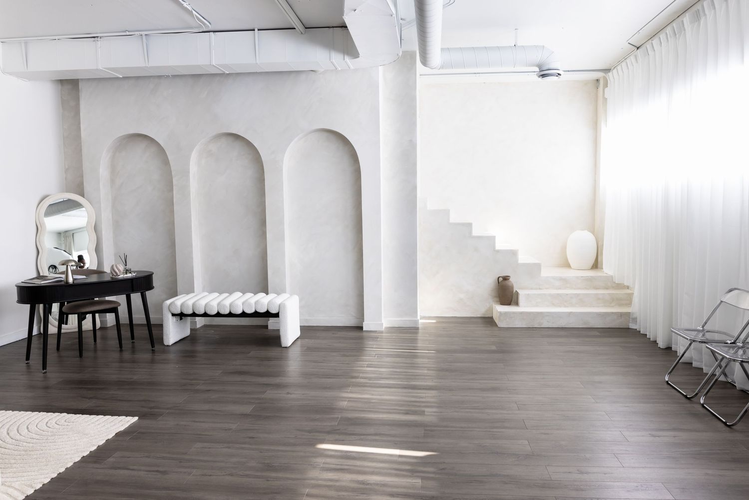 Minimalist white studio space with arched walls, dark hardwood floors, vanity mirror, bench, and white staircase.