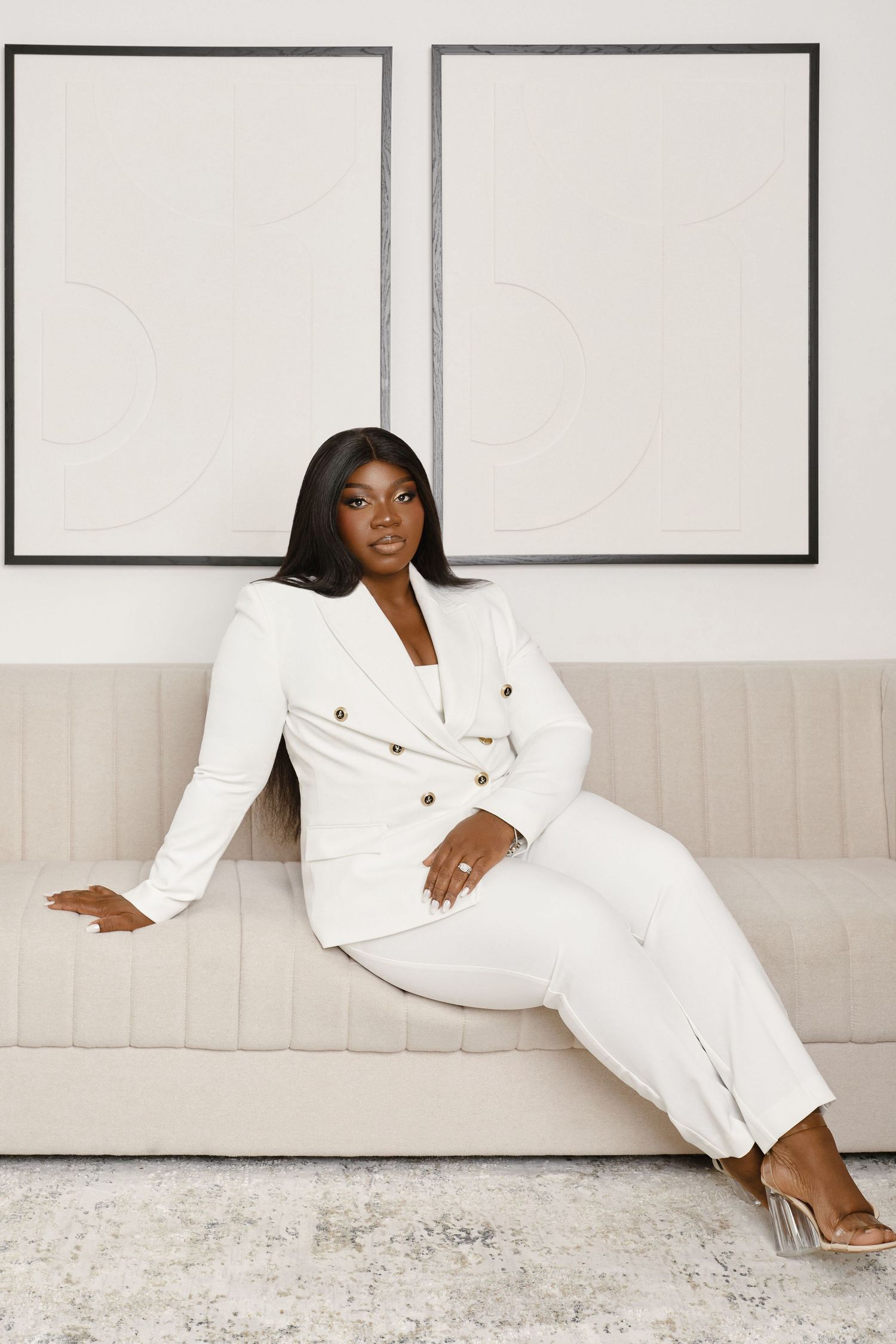 Fashion model in white suit lounges elegantly against wall in minimalist studio setting.