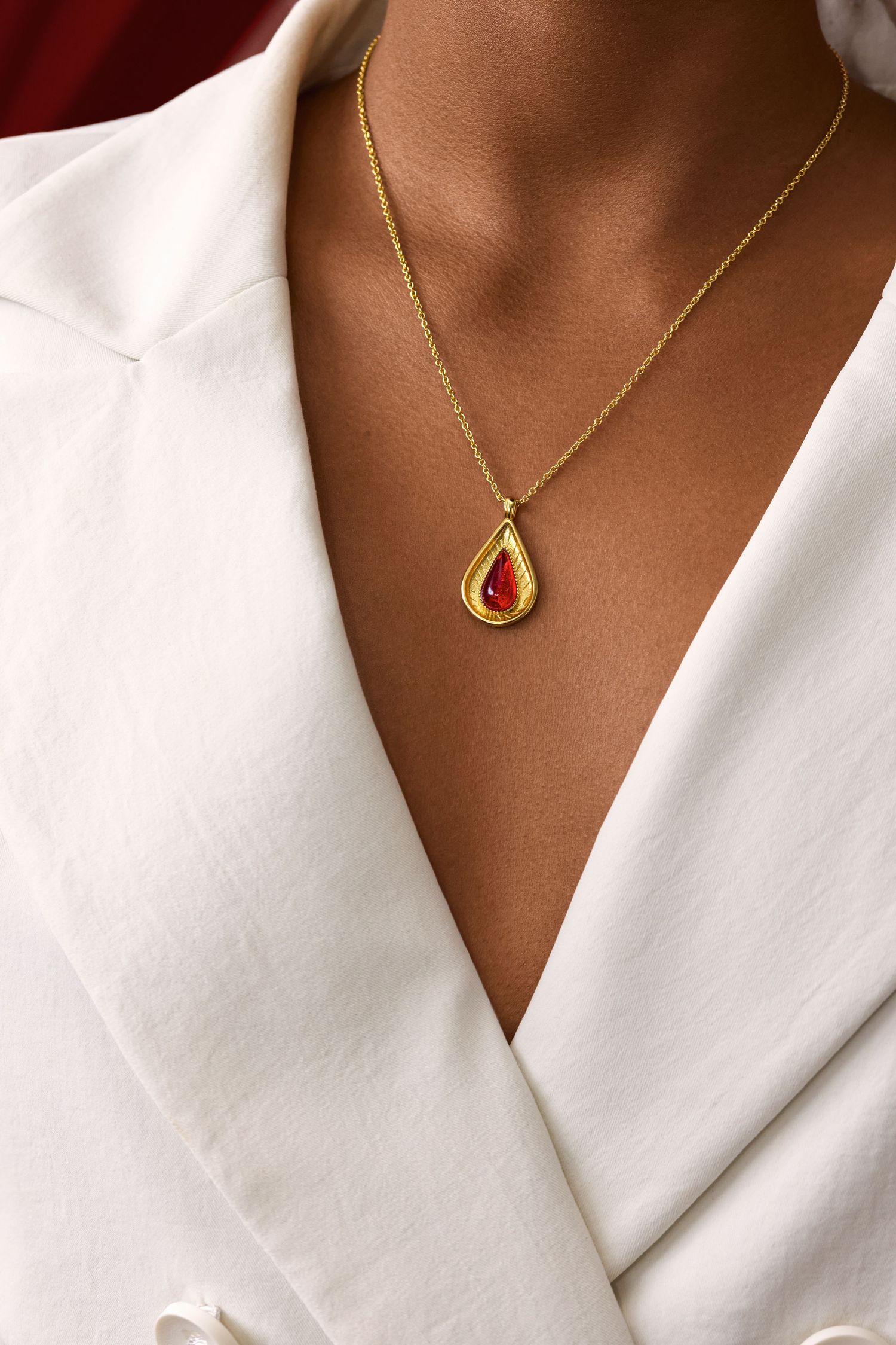 Gold teardrop pendant necklace with red stone worn against white blazer on tan skin.