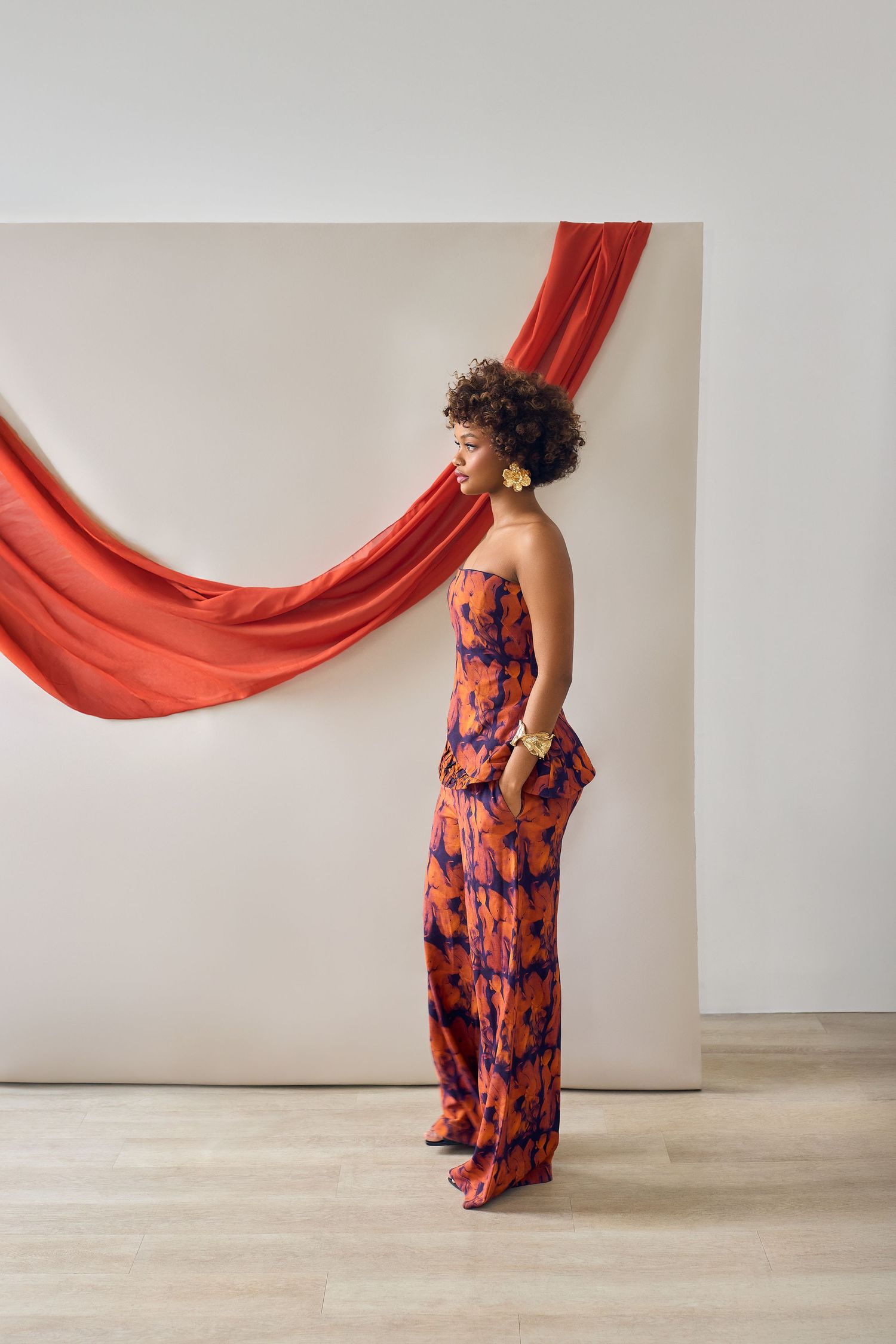 Woman in strapless orange floral jumpsuit poses elegantly before white wall with draped red fabric backdrop.