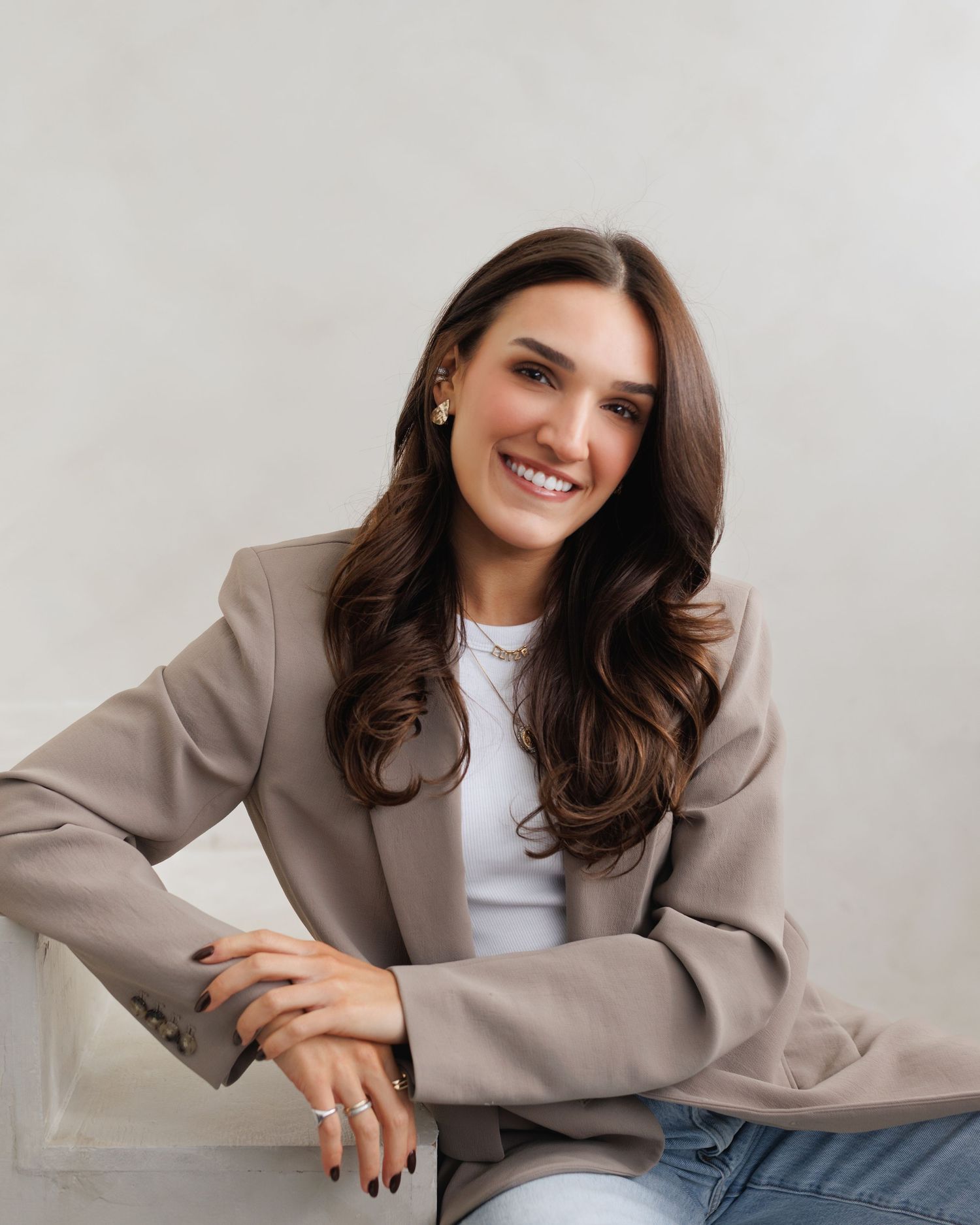 Smiling brunette woman in beige blazer and white top poses casually in a bright studio setting.