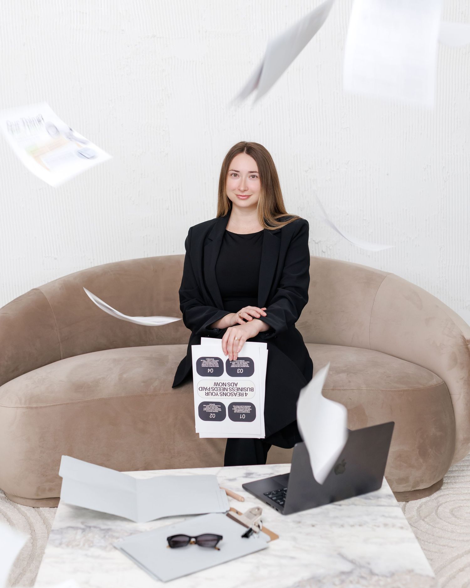 Young professional woman in black blazer sitting on beige sofa holding documents with laptop and papers flying around.
