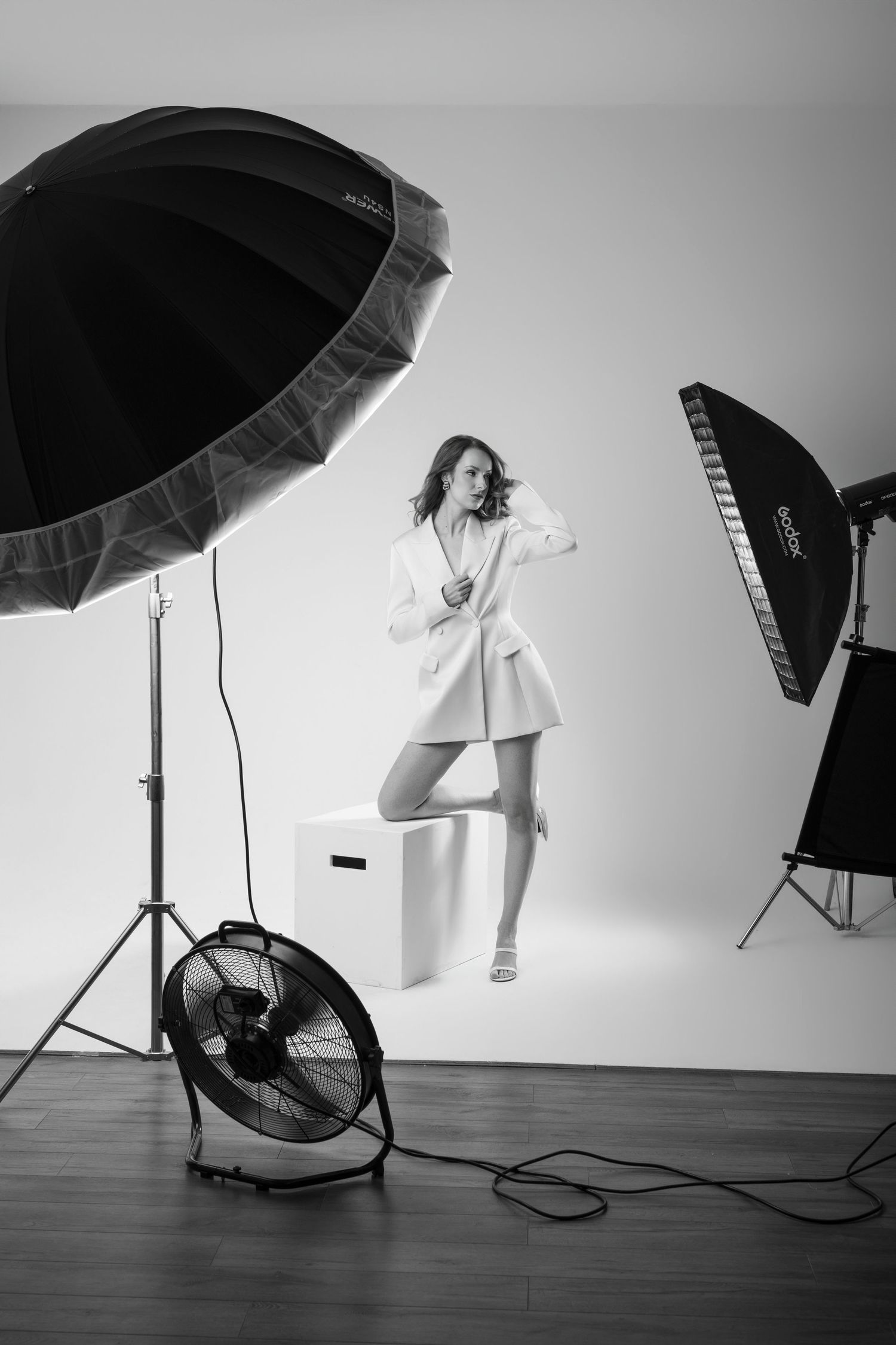 Black and white studio photo of a woman in a white dress posing with flash umbrellas and a fan.