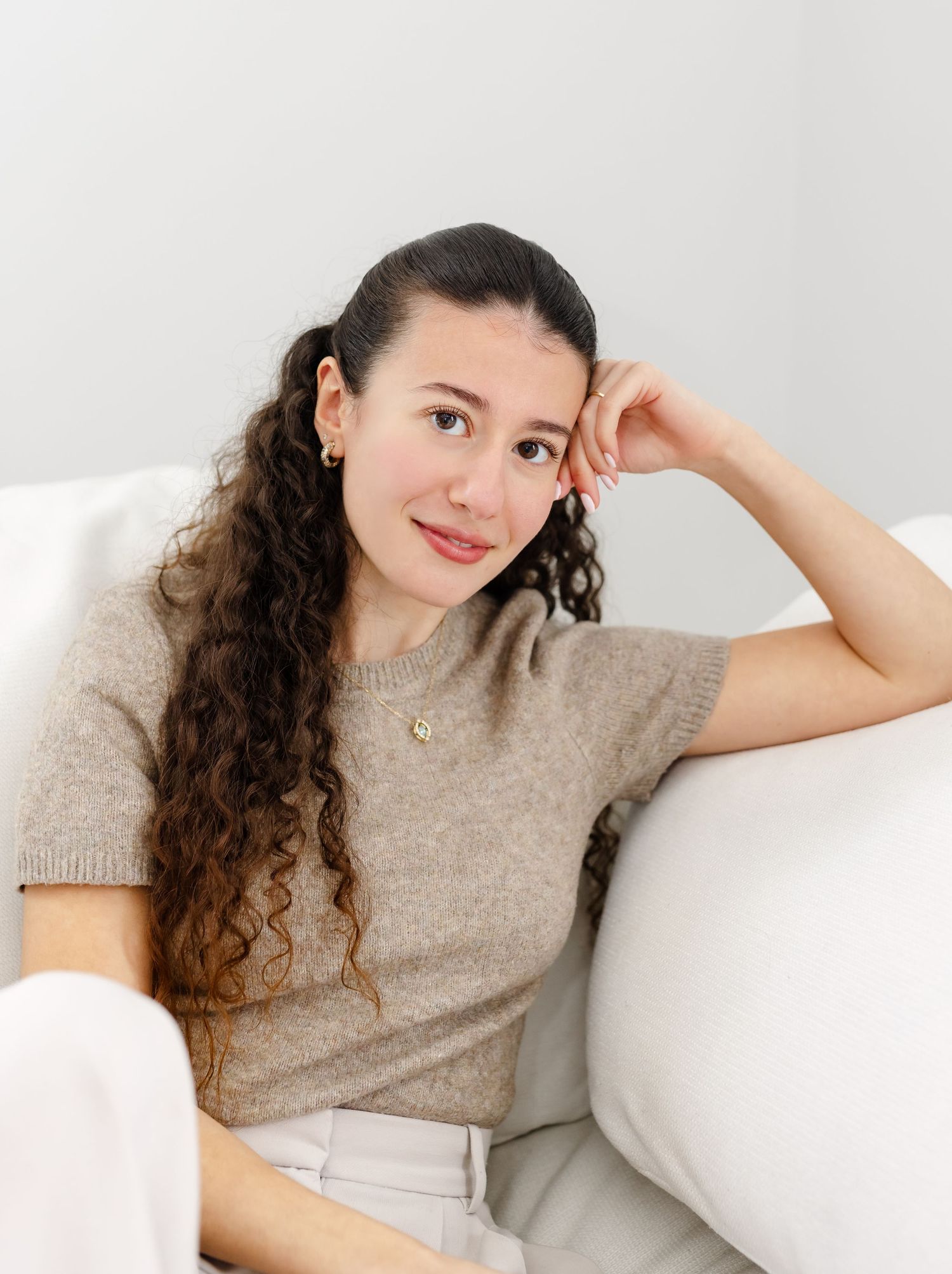 A person with long curly brown hair sits relaxed on a white couch wearing a beige sweater and white pants.