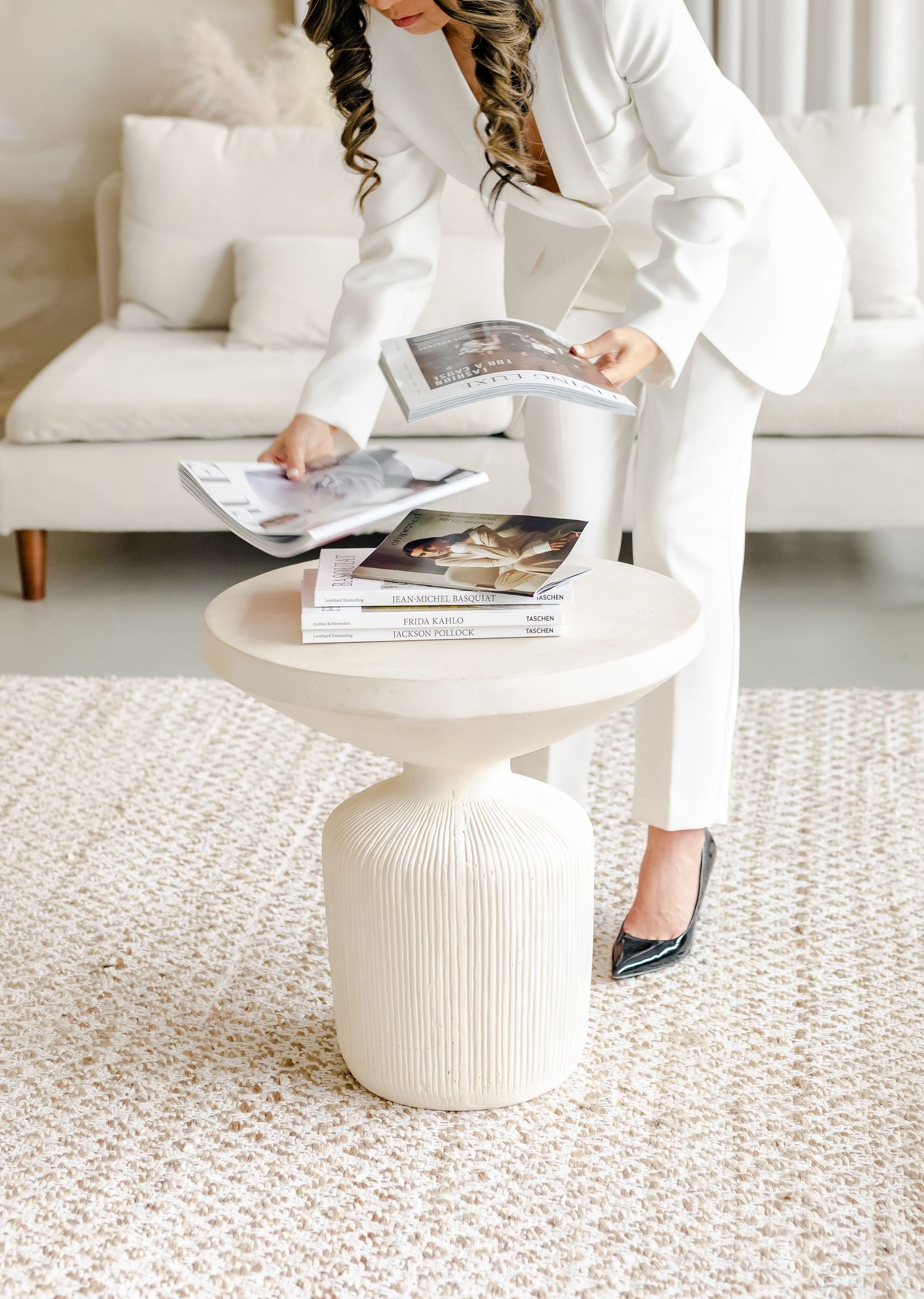 Elegant all-white living room interior with person arranging magazines on textured ceramic side table over neutral carpet.