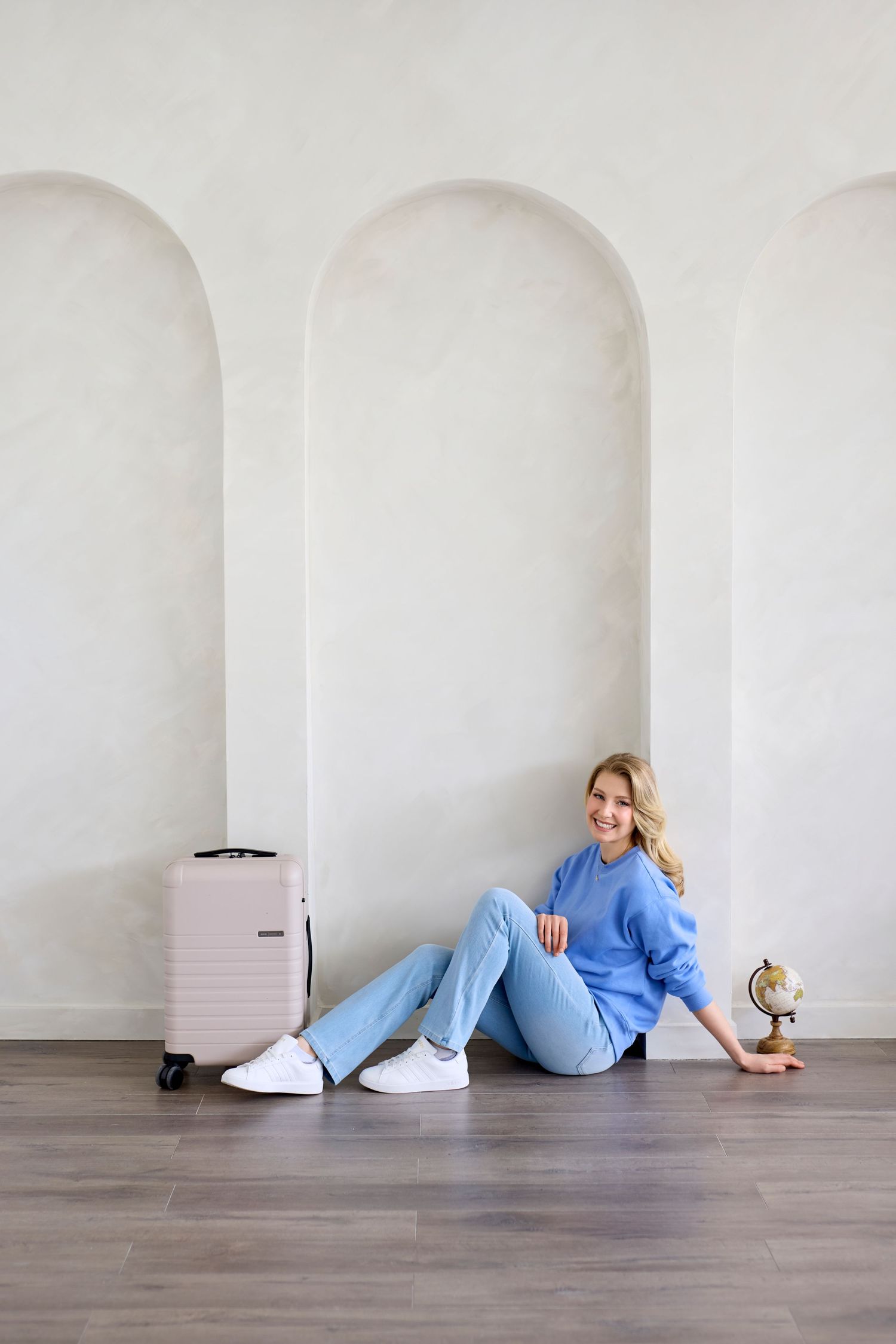 A relaxed figure in a blue sweater and jeans sits on the floor next to pink luggage and small globe in a white archway.
