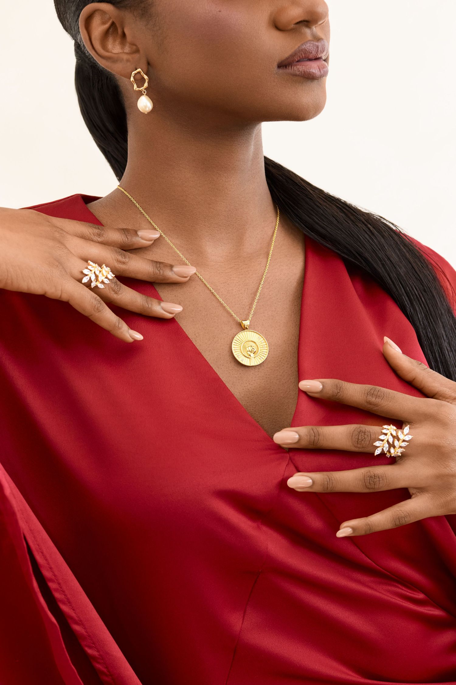 Close up woman in red blouse displaying gold jewelry on fingers, neck, and ears against white background.