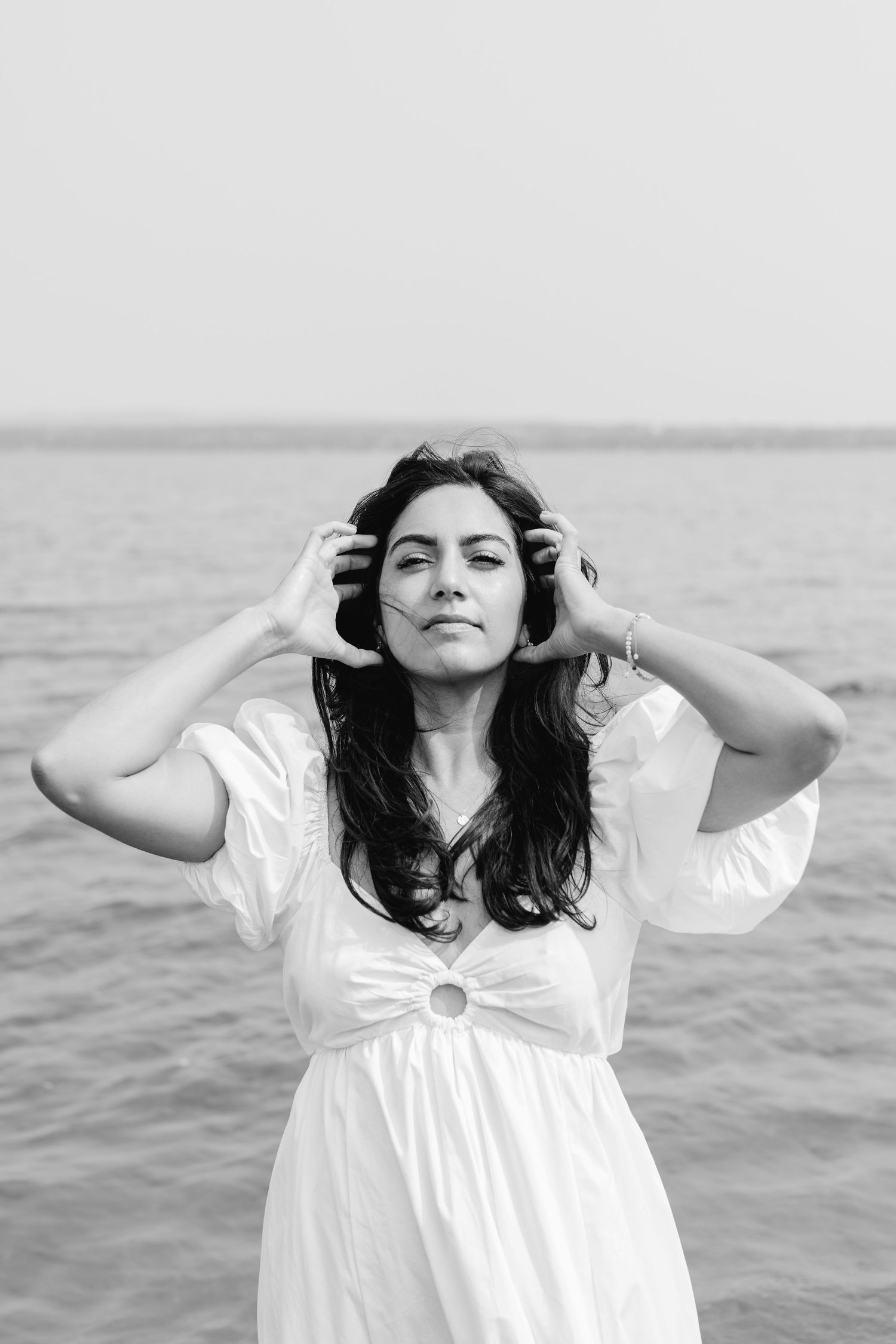 A person in a white dress stands by the water with hands in their hair on an overcast day.