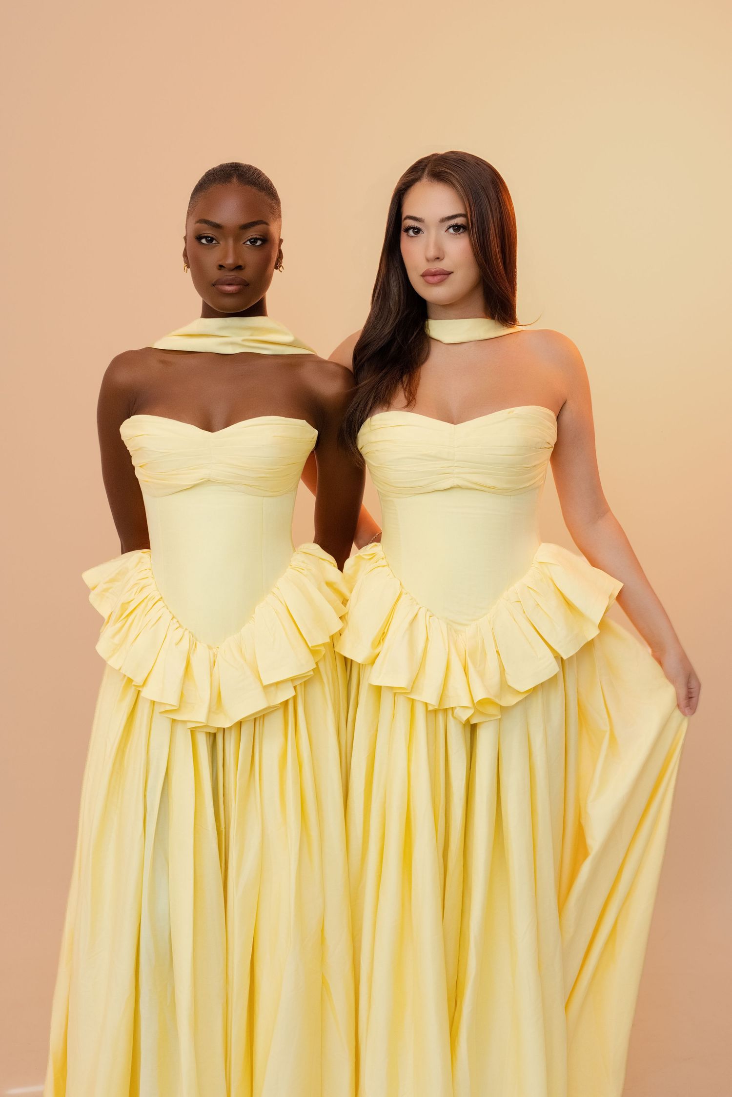 Two women in matching yellow ruffled strapless gowns with neck scarves pose against a beige background.