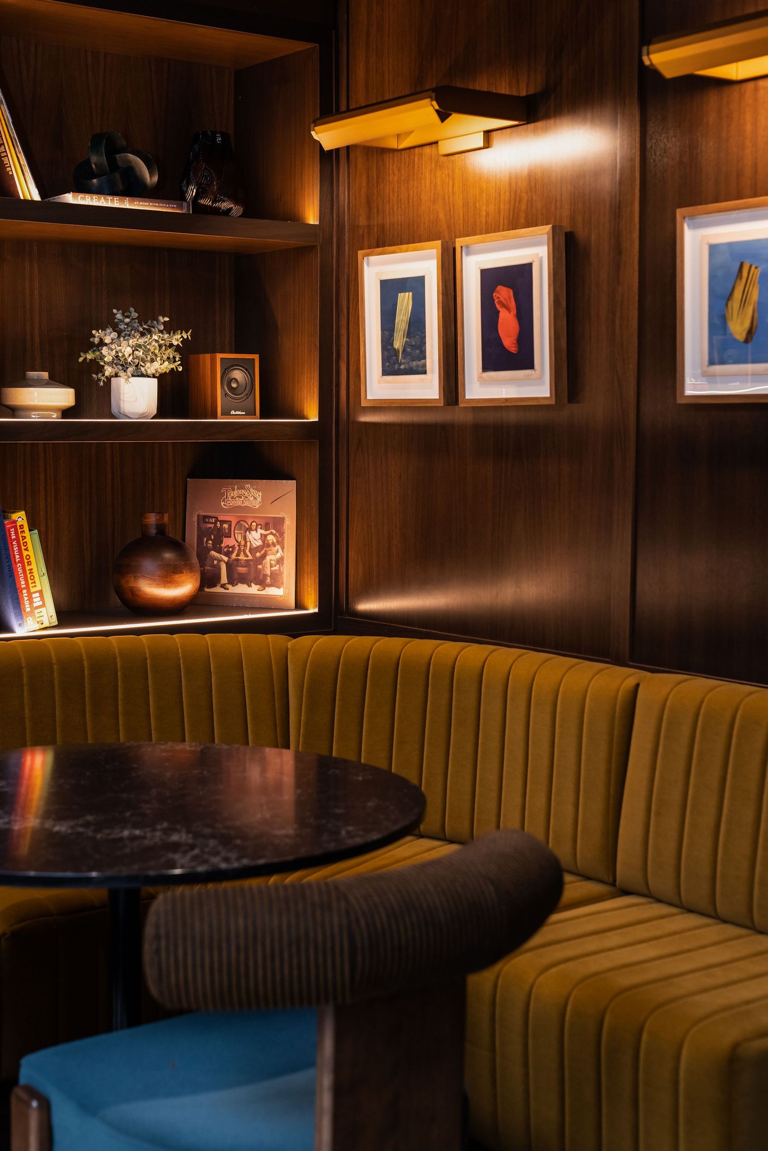 Cozy mid-century modern restaurant booth with olive velvet seating, dark wood paneling, framed art, and warm ambient lighting.