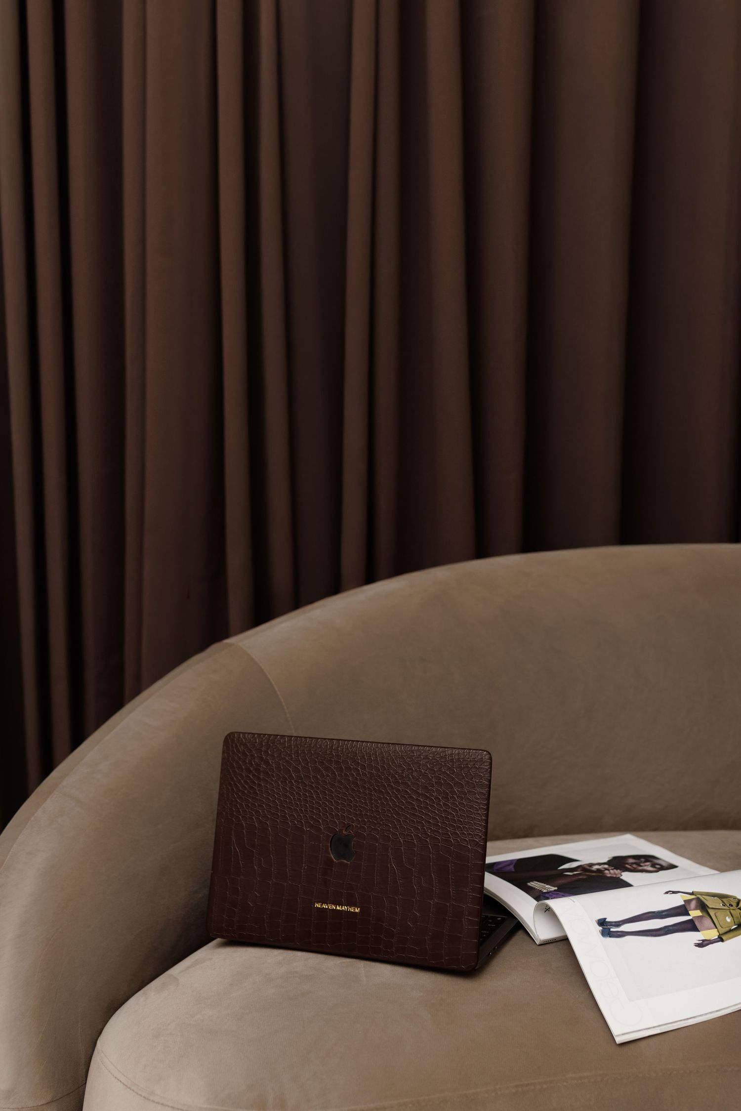 Dark brown crocodile leather clutch and fashion magazine on a beige curved sofa against rich brown curtains.