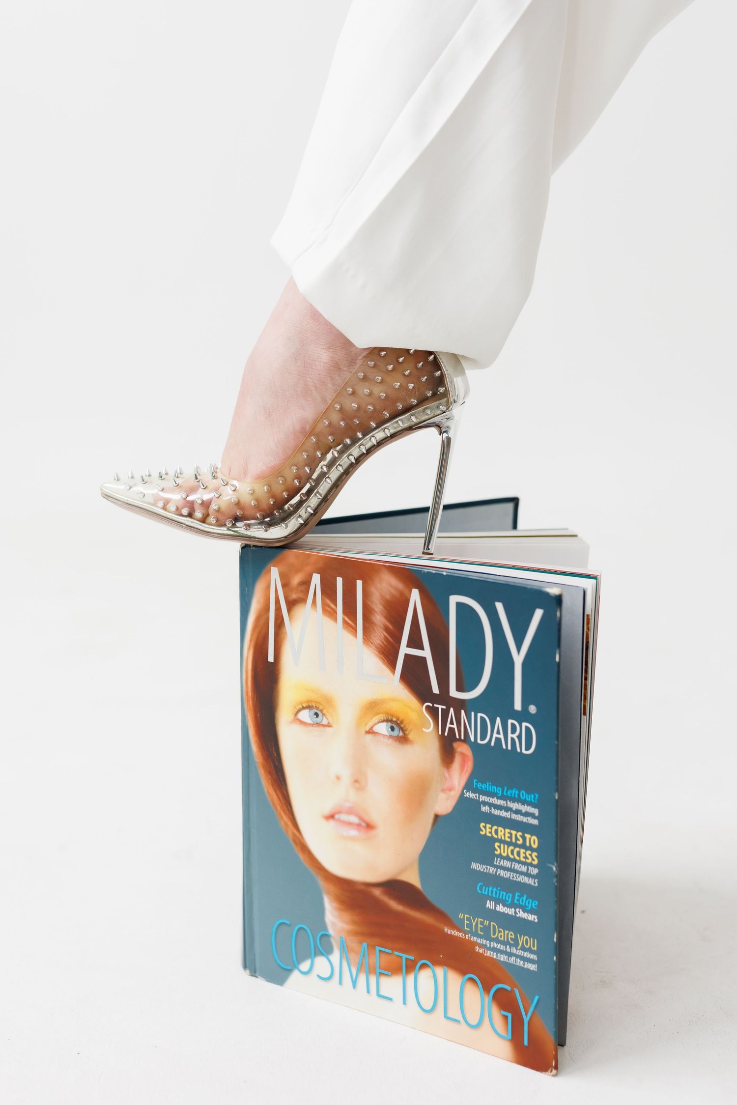 Studded high heel stepping on fashion magazines against white background.
