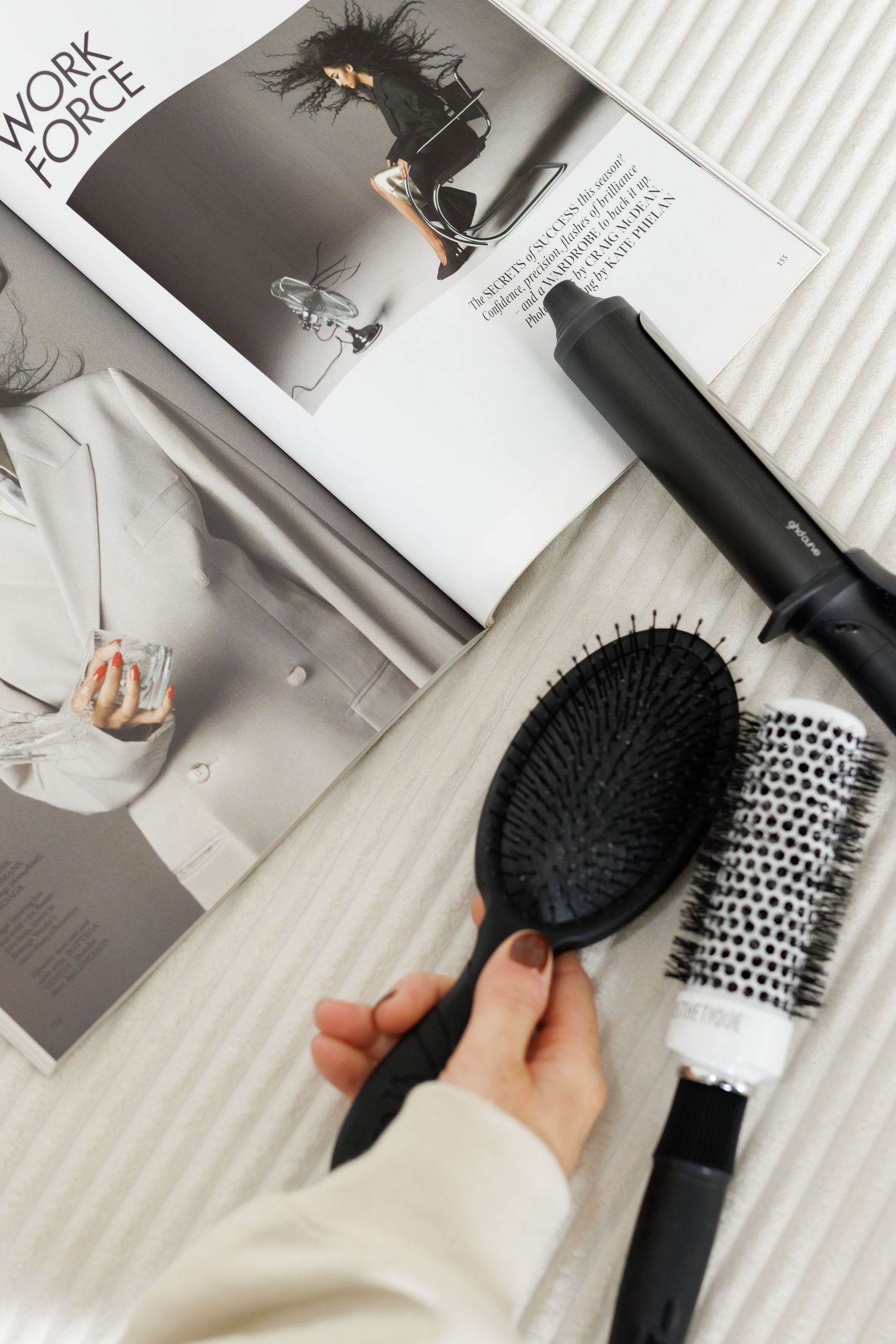 Styling tools and magazine layout on white surface with hair brushes and curling iron.