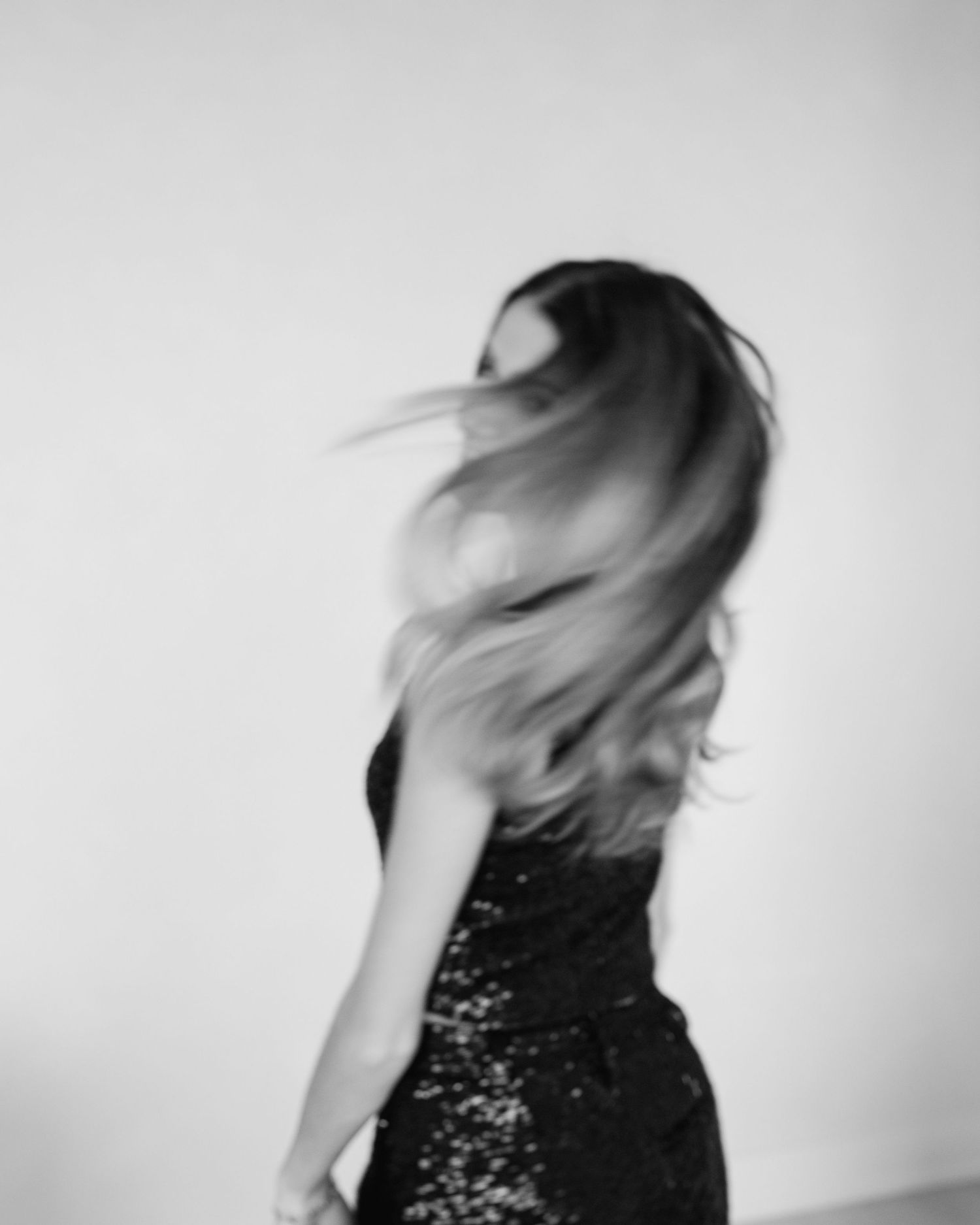 Black and white artistic portrait of someone in sequined dress whipping their hair in dynamic motion against light background.