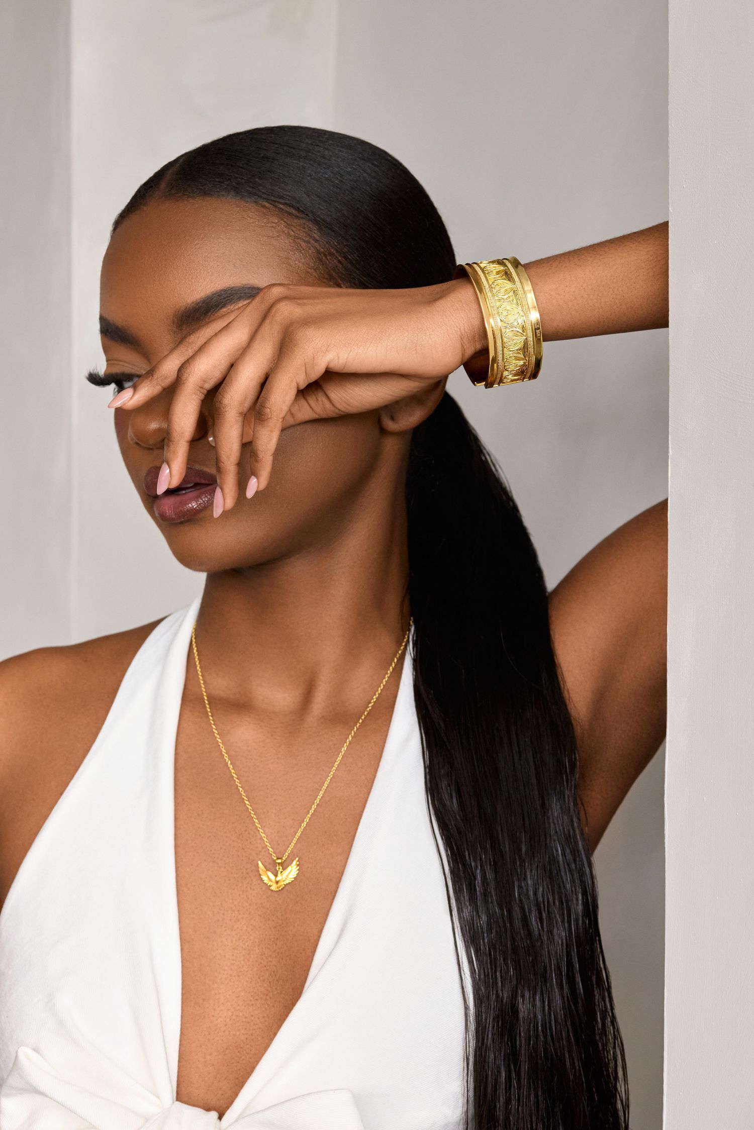 Black woman in white halter top wearing gold bangles and necklace, posing with hand over face against neutral background.