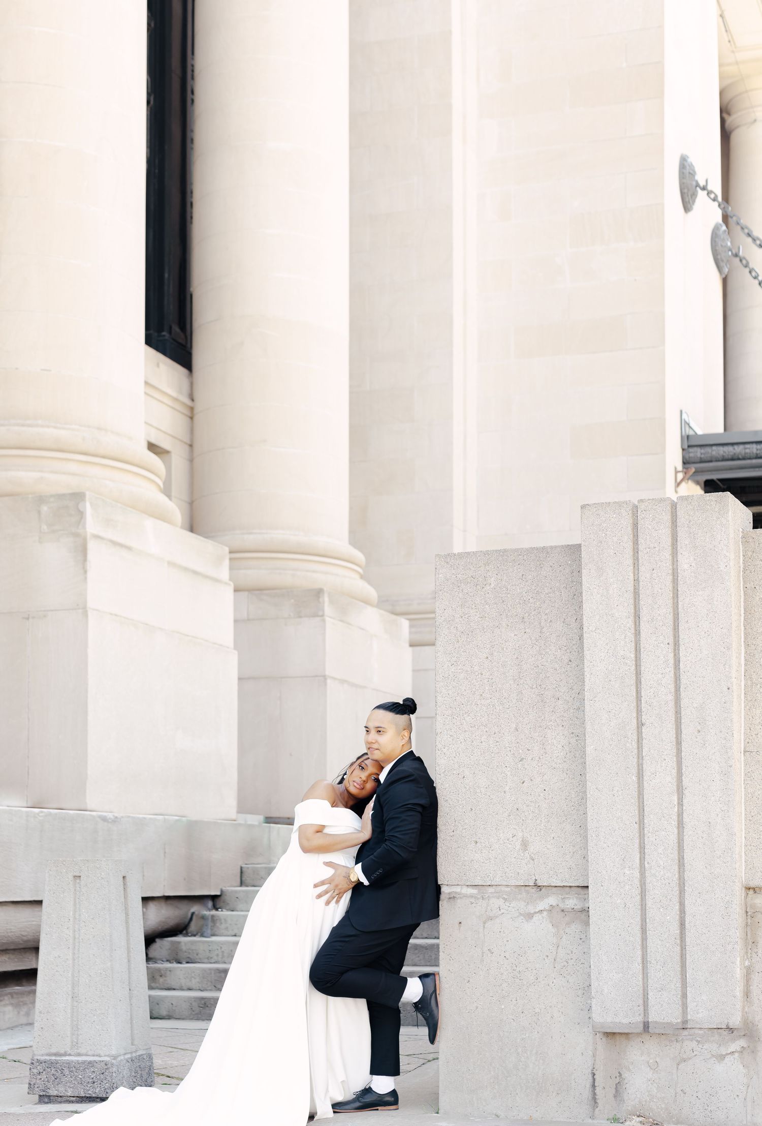 A couple in wedding attire playfully pose between white classical columns of a grand building.