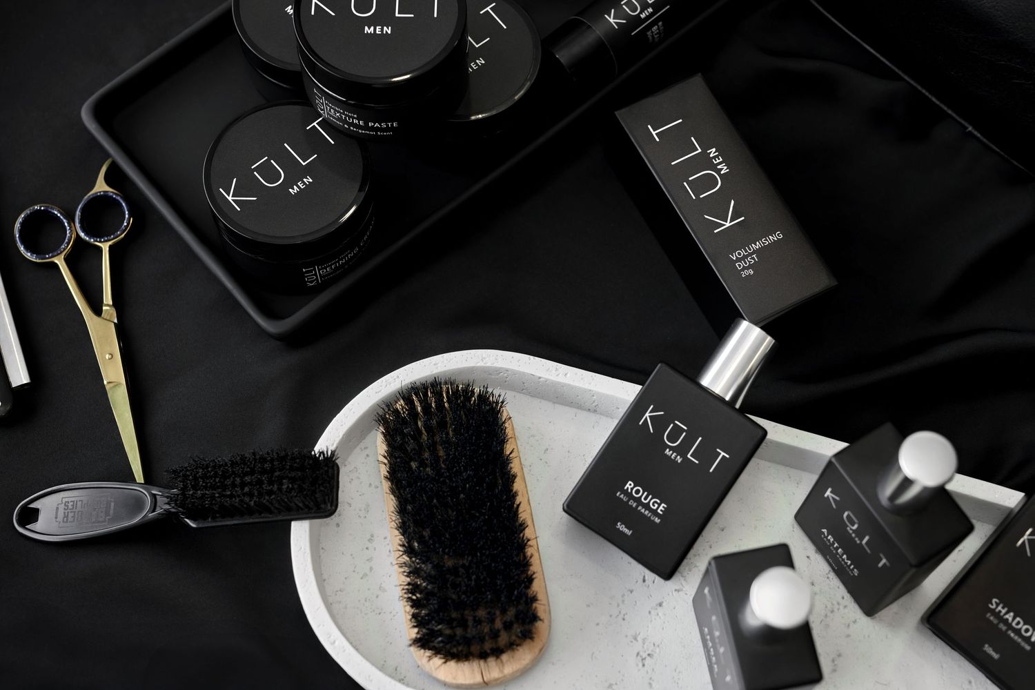 Black and white flatlay of luxury hair care products, grooming tools and accessories on dark surface with brush and scissors.