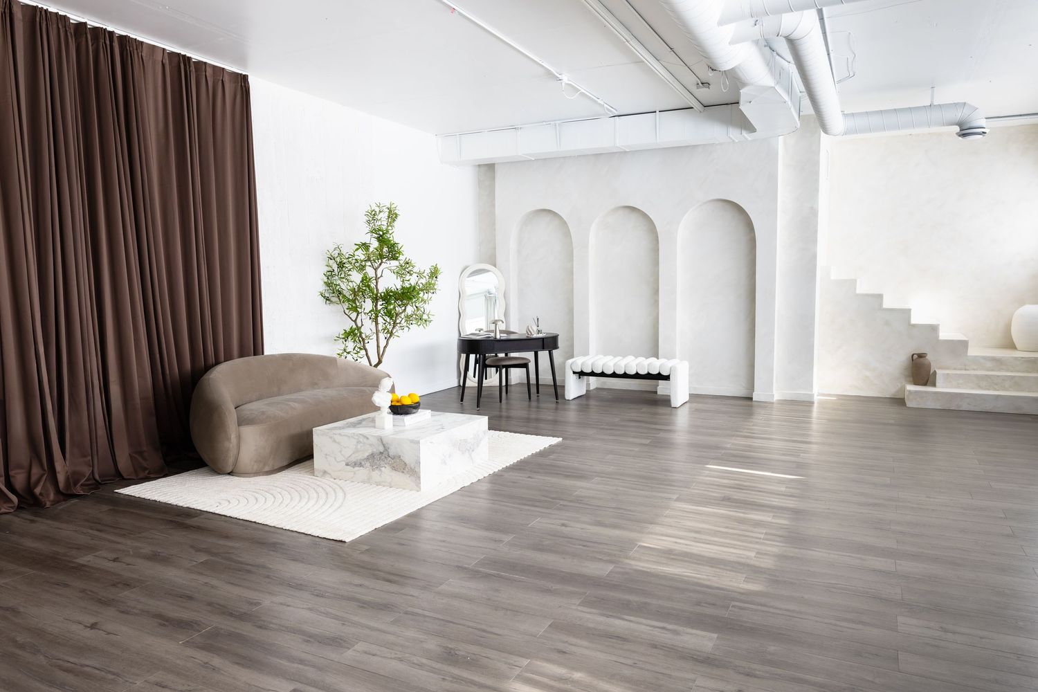 Modern minimalist loft with gray hardwood floors, arched walls, beige chair, marble coffee table, and brown curtains.