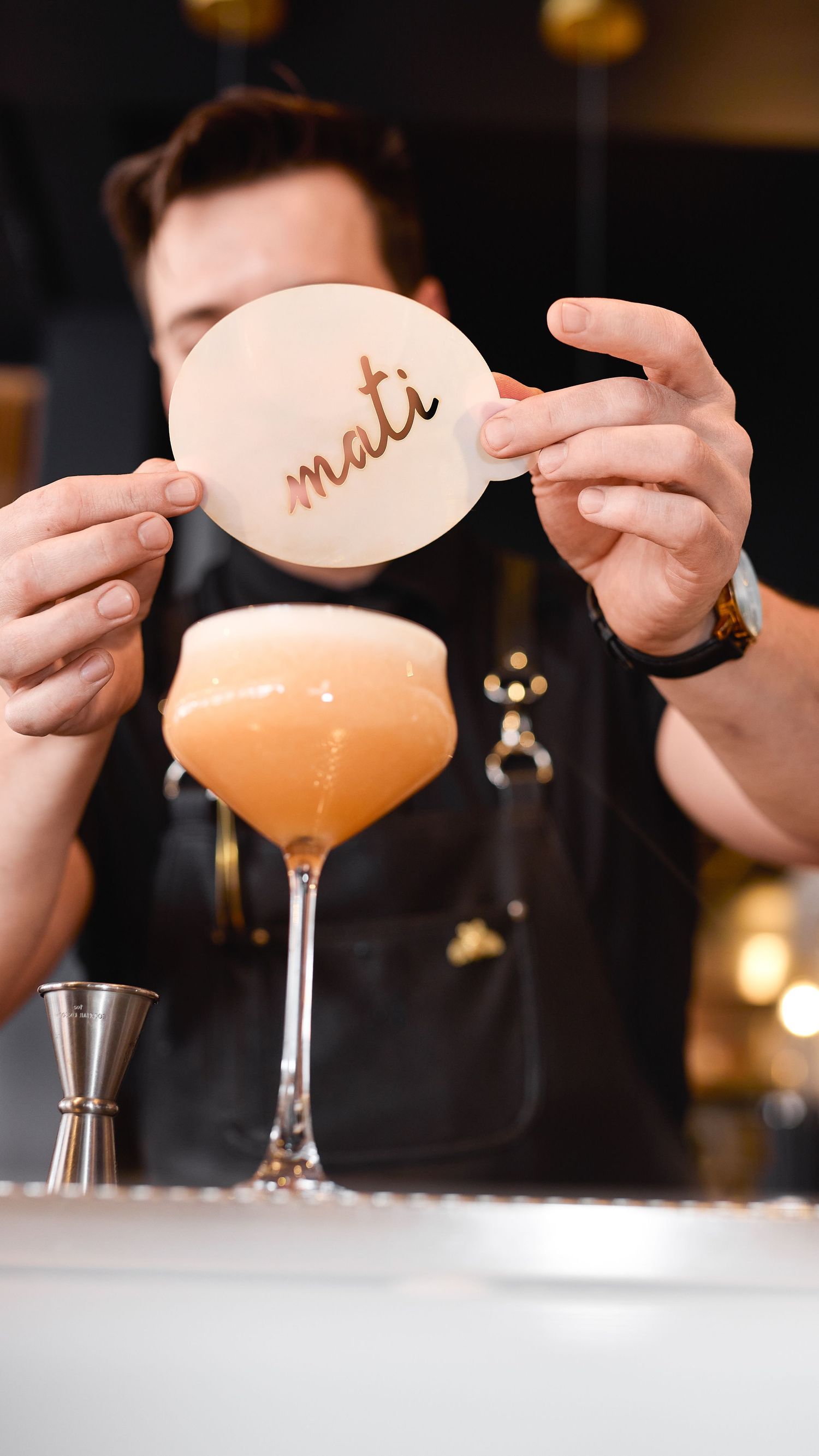 Hands placing a decorative garnish on top of a pink cocktail in a coupe glass at a dimly lit bar.