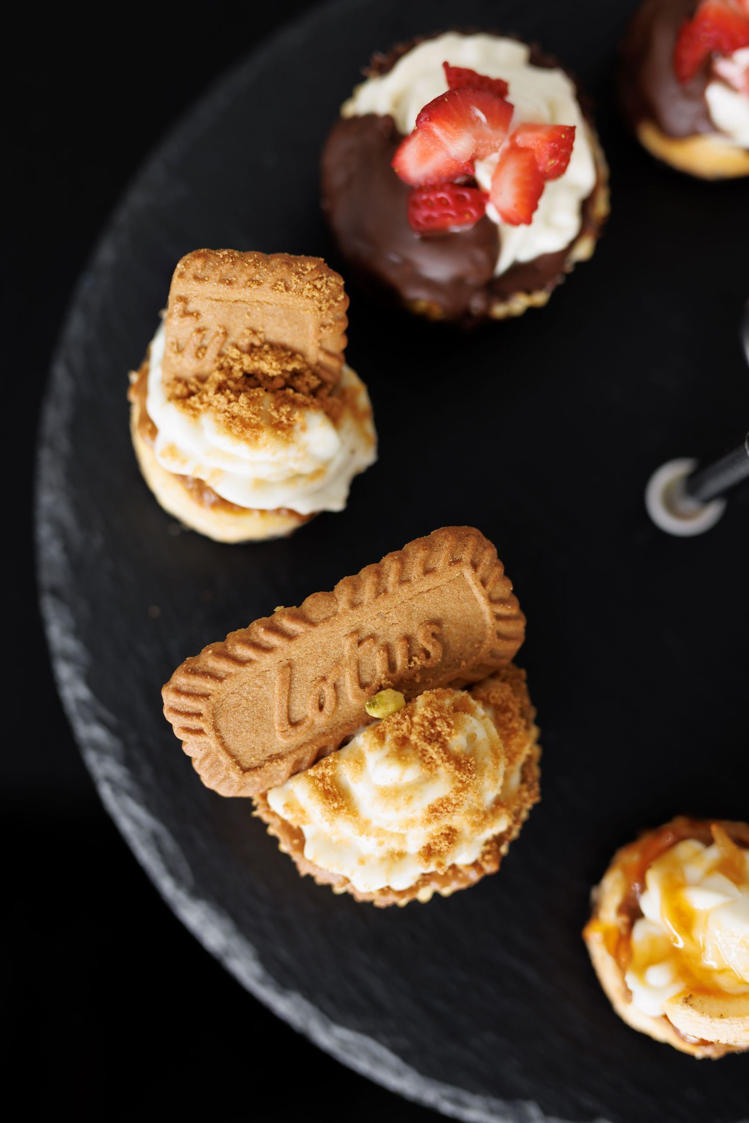 Assorted gourmet dessert bites including chocolate-dipped treats with strawberries and Lotus cookies on a black slate plate.