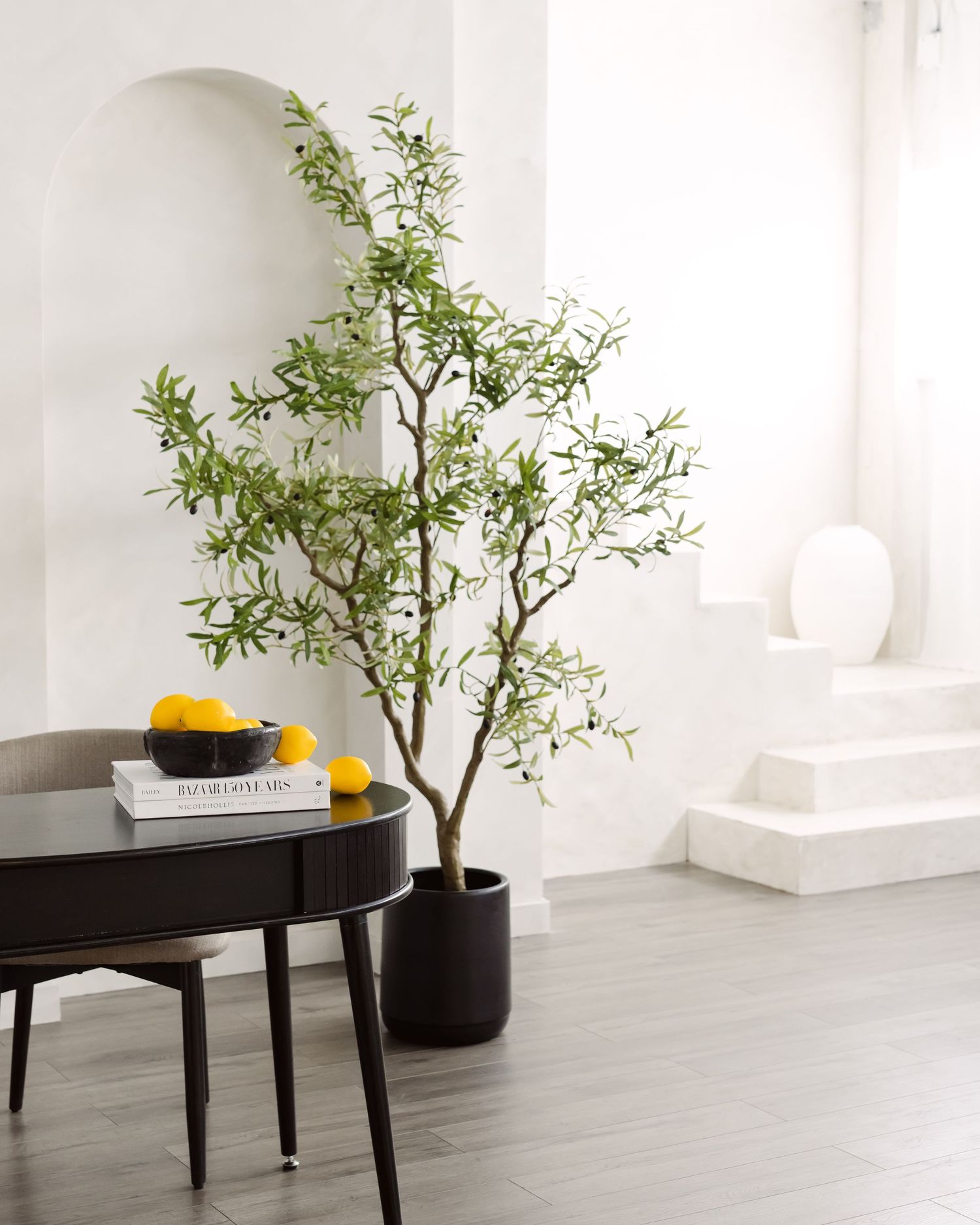 Tall artificial olive tree in black pot beside dark round coffee table with lemons, in bright white modern living room.