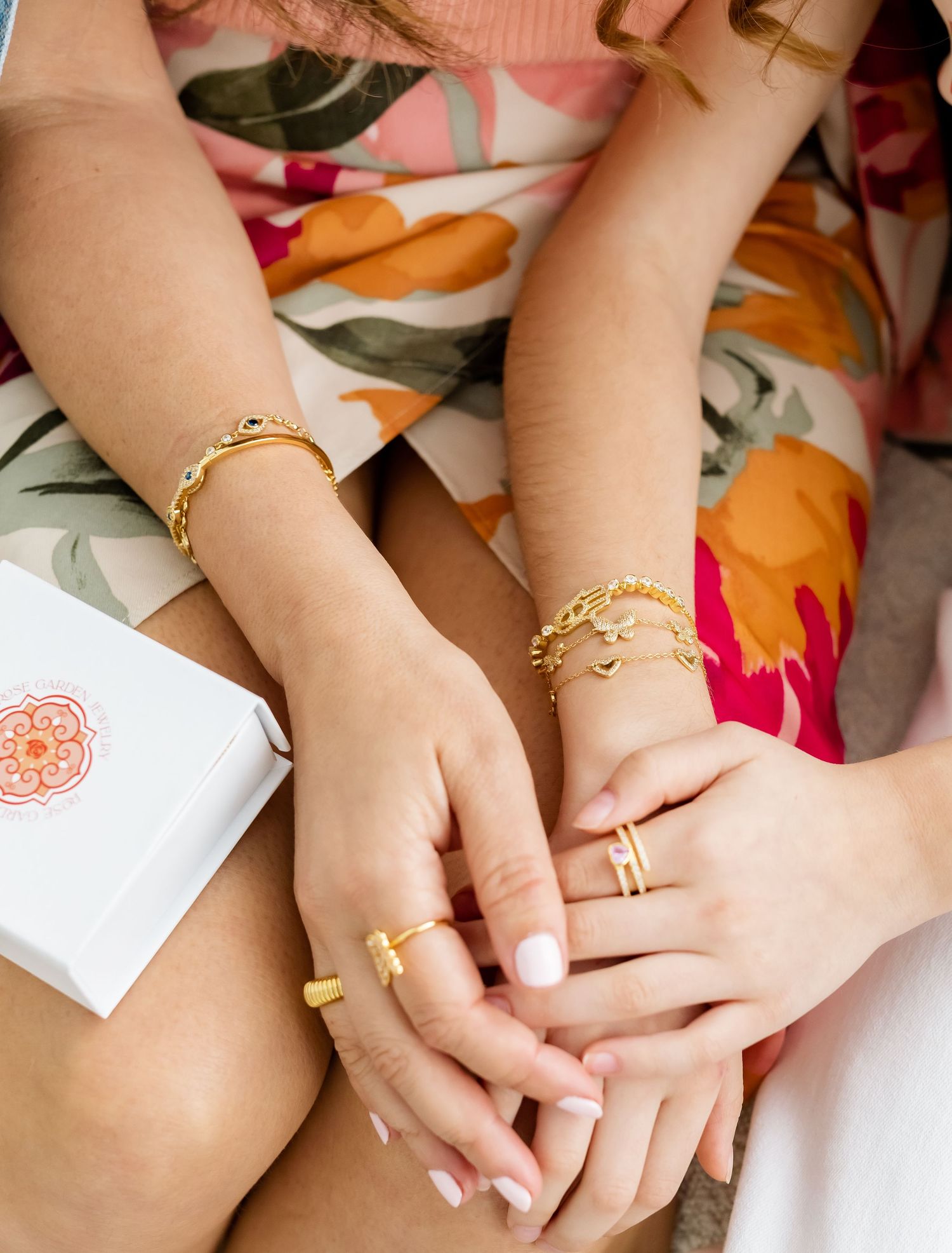 Two women wearing layered gold bracelets and rings with white manicured nails, sitting together in floral outfits.