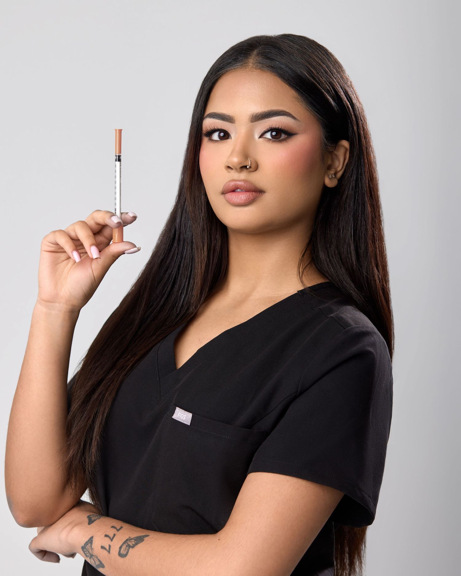 A female medical professional in black scrubs holds a syringe, showcasing her tattoo and long dark hair.