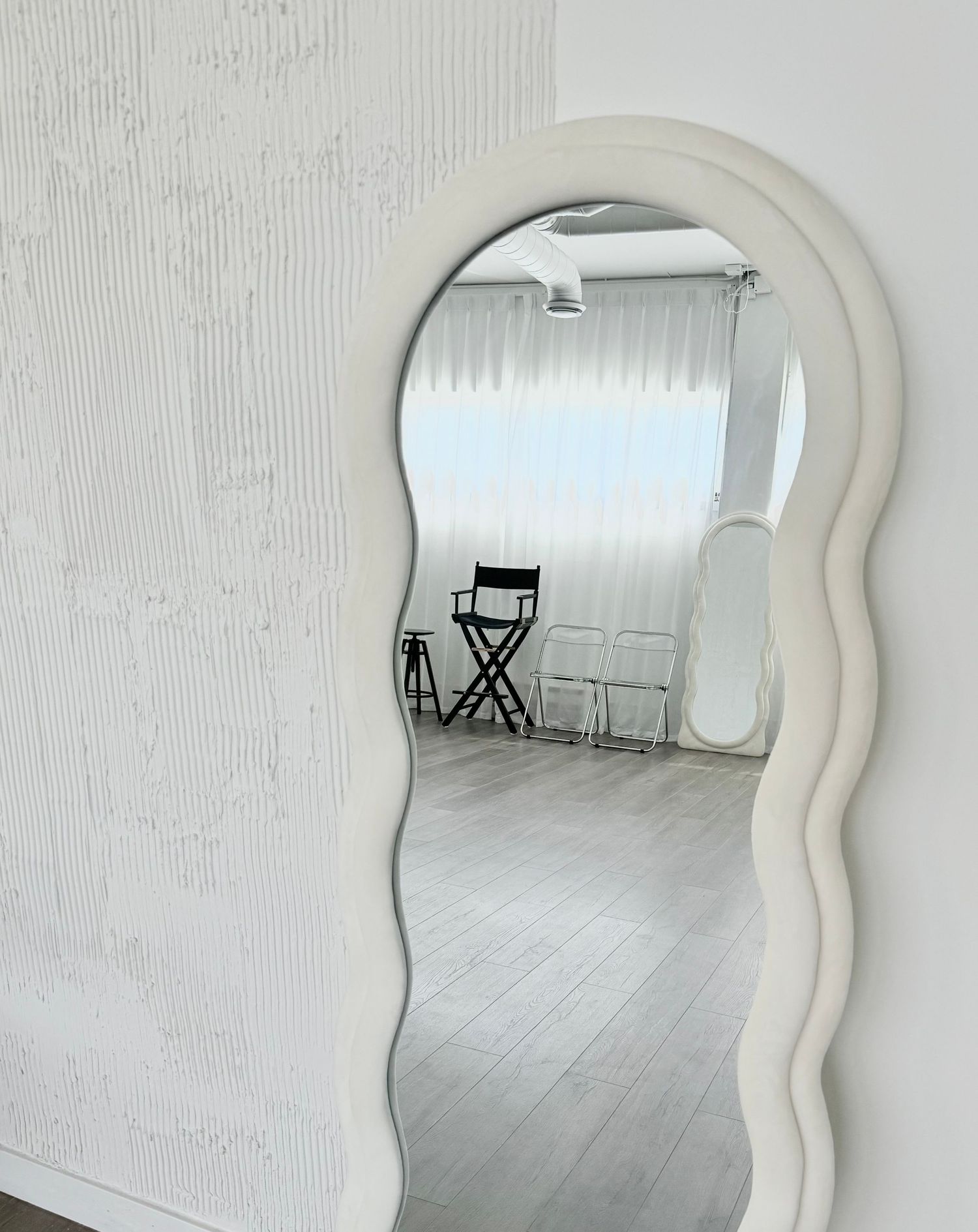 A curvy white decorative mirror frames a view of a minimalist white room with gray wood flooring and natural lighting.