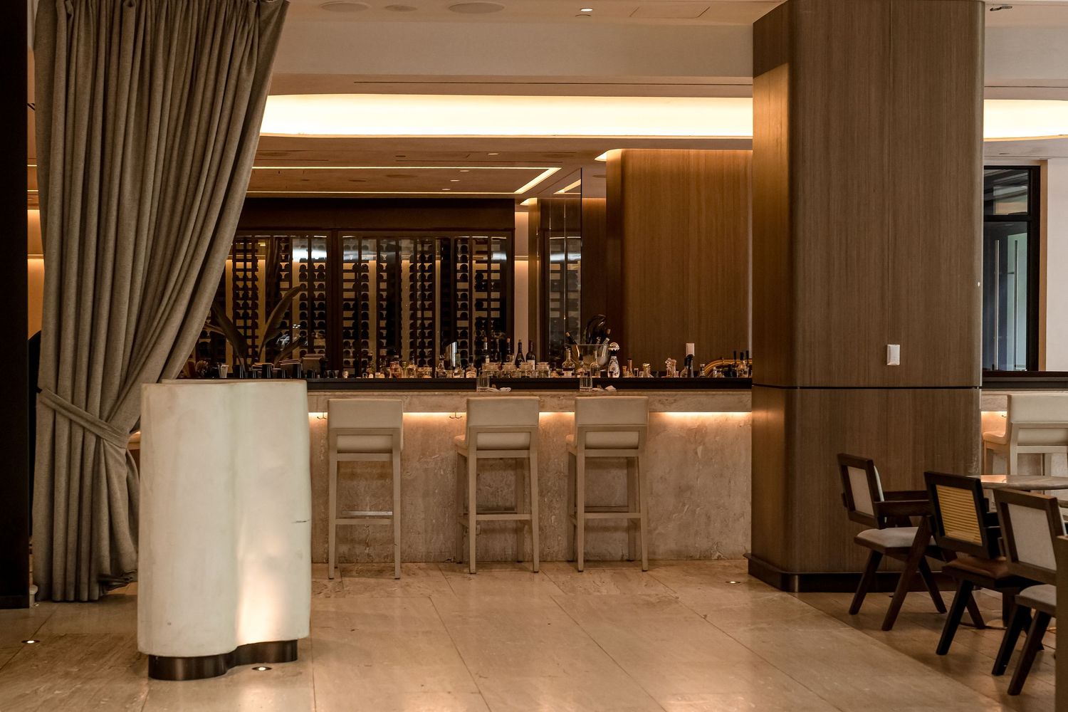 Elegant hotel bar with wood paneling, wine display, marble counter, and bar stools in a warmly lit upscale lounge.