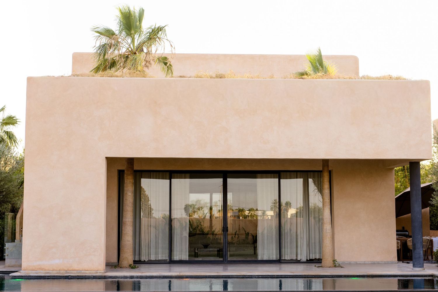 Modern Moroccan villa with terracotta walls, sliding glass doors, and palm trees against a clear sky.