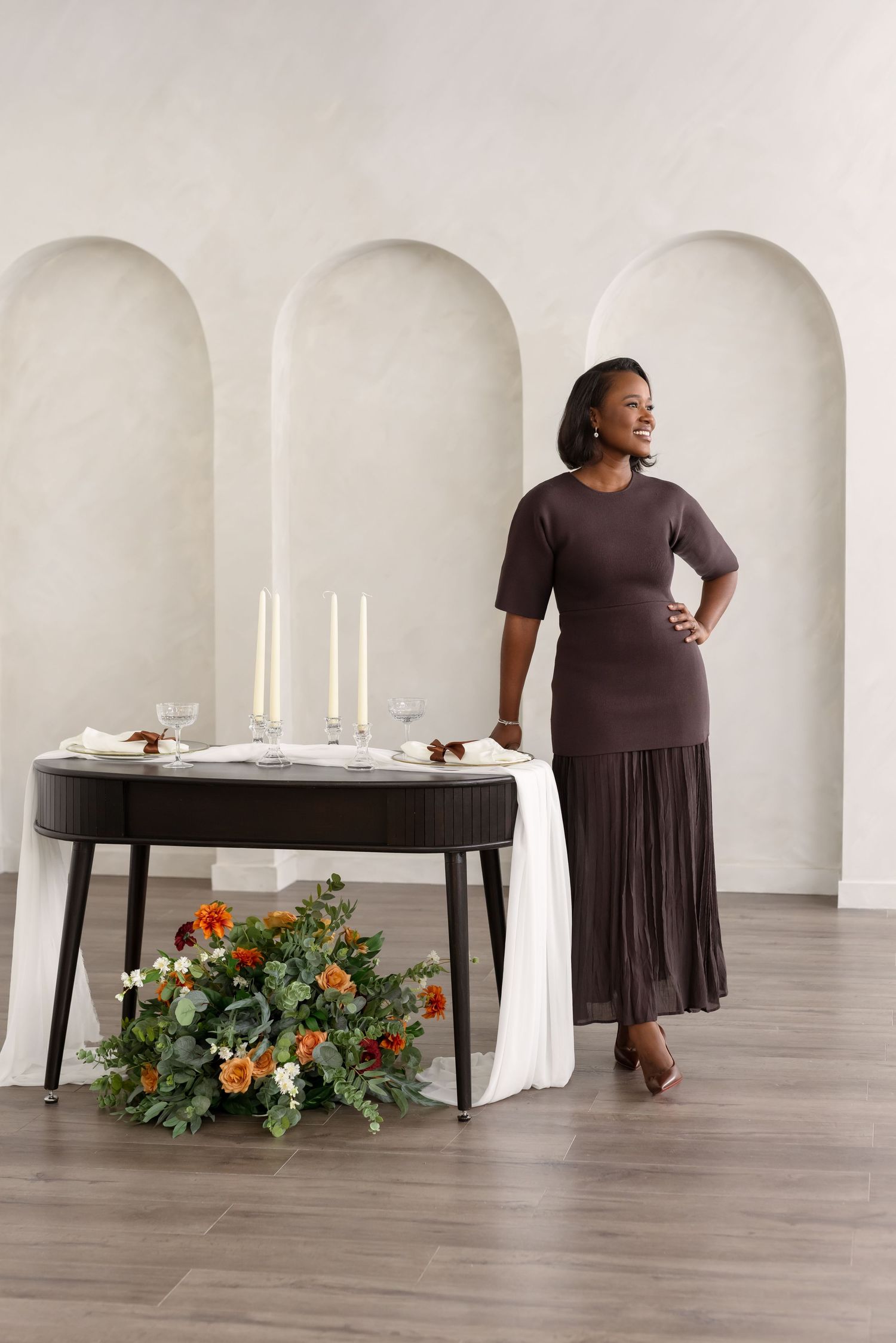 A person in a brown dress poses in a minimalist white room with a table decorated with candles and floral arrangements.