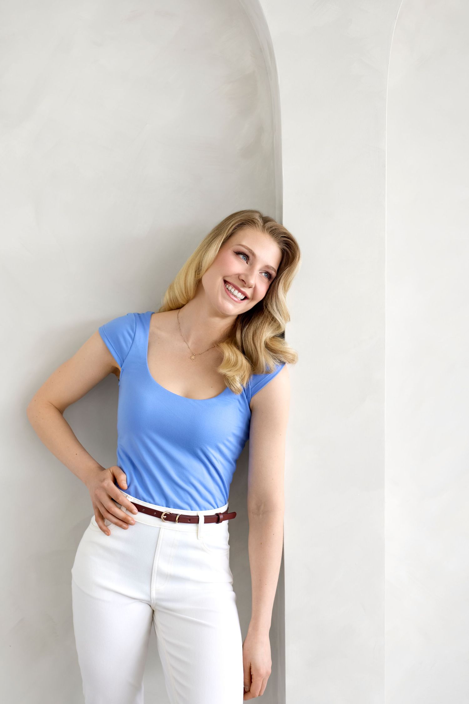 Casual summer look with blue fitted top and white pants against light wall.