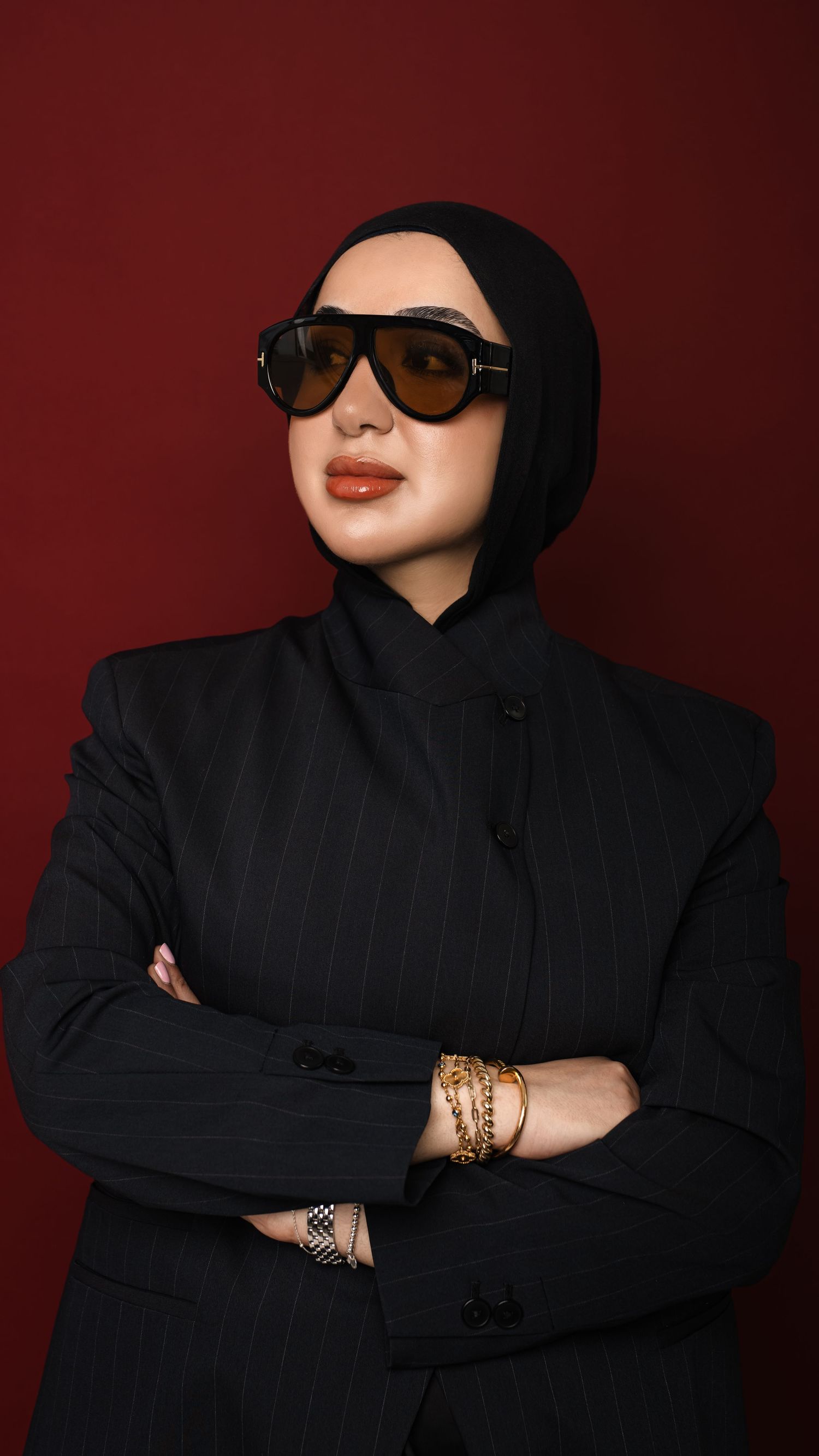 Woman in black hijab and pinstripe blazer wearing oversized sunglasses and gold bracelets poses confidently against red background.