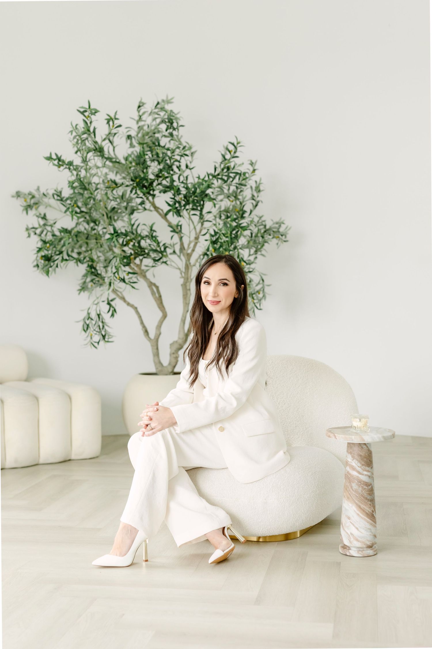 Woman in white suit sitting elegantly in a modern white room with a green tree and marble side table.