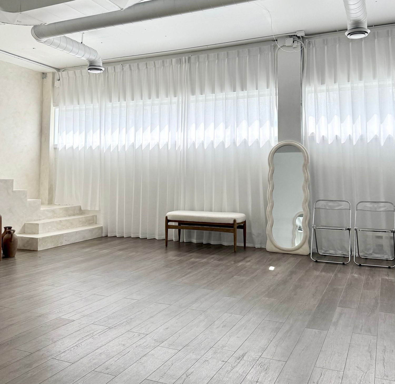 Minimalist dance studio interior with white curtains, wood flooring, full-length mirror and folding chairs along the wall.