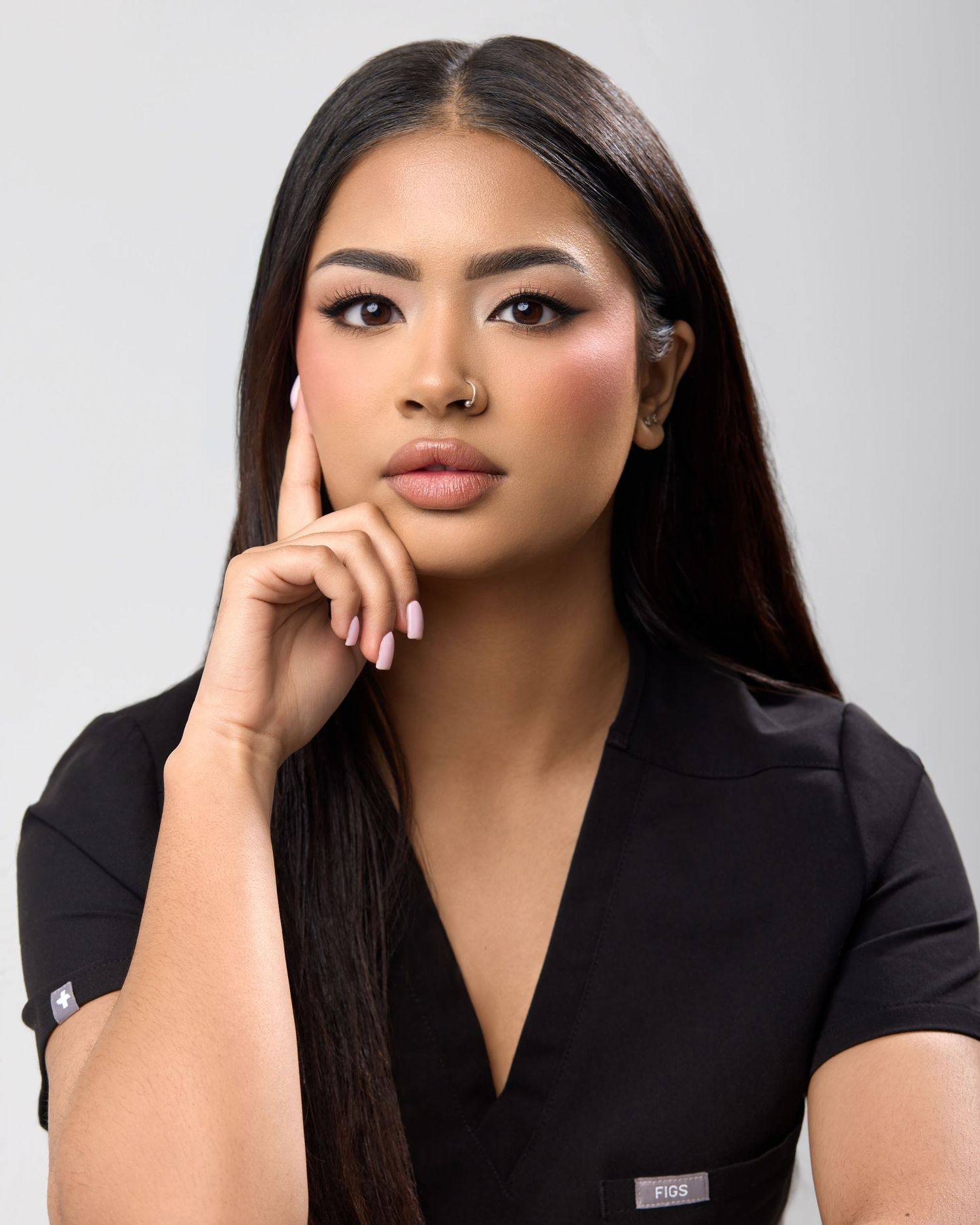 Young woman in black scrubs poses confidently with hand on chin, wearing glamorous makeup and long dark hair.