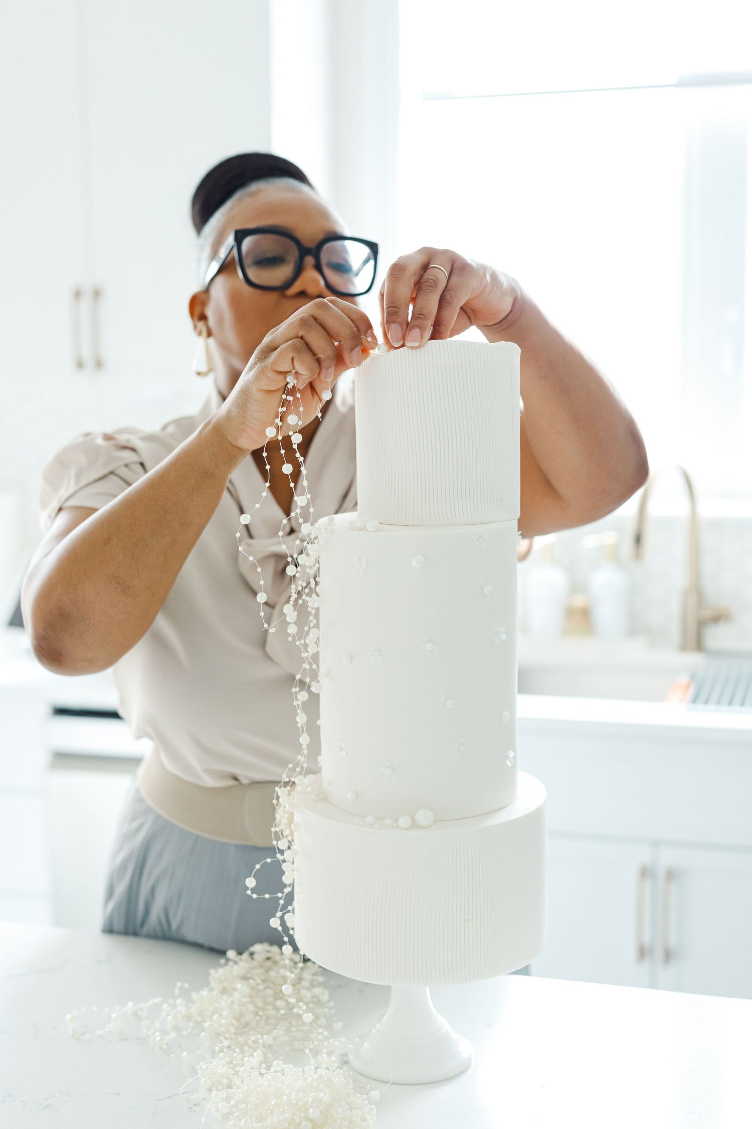 A pastry chef in glasses decorates a three-tiered white cake with sprinkles in a bright kitchen setting.