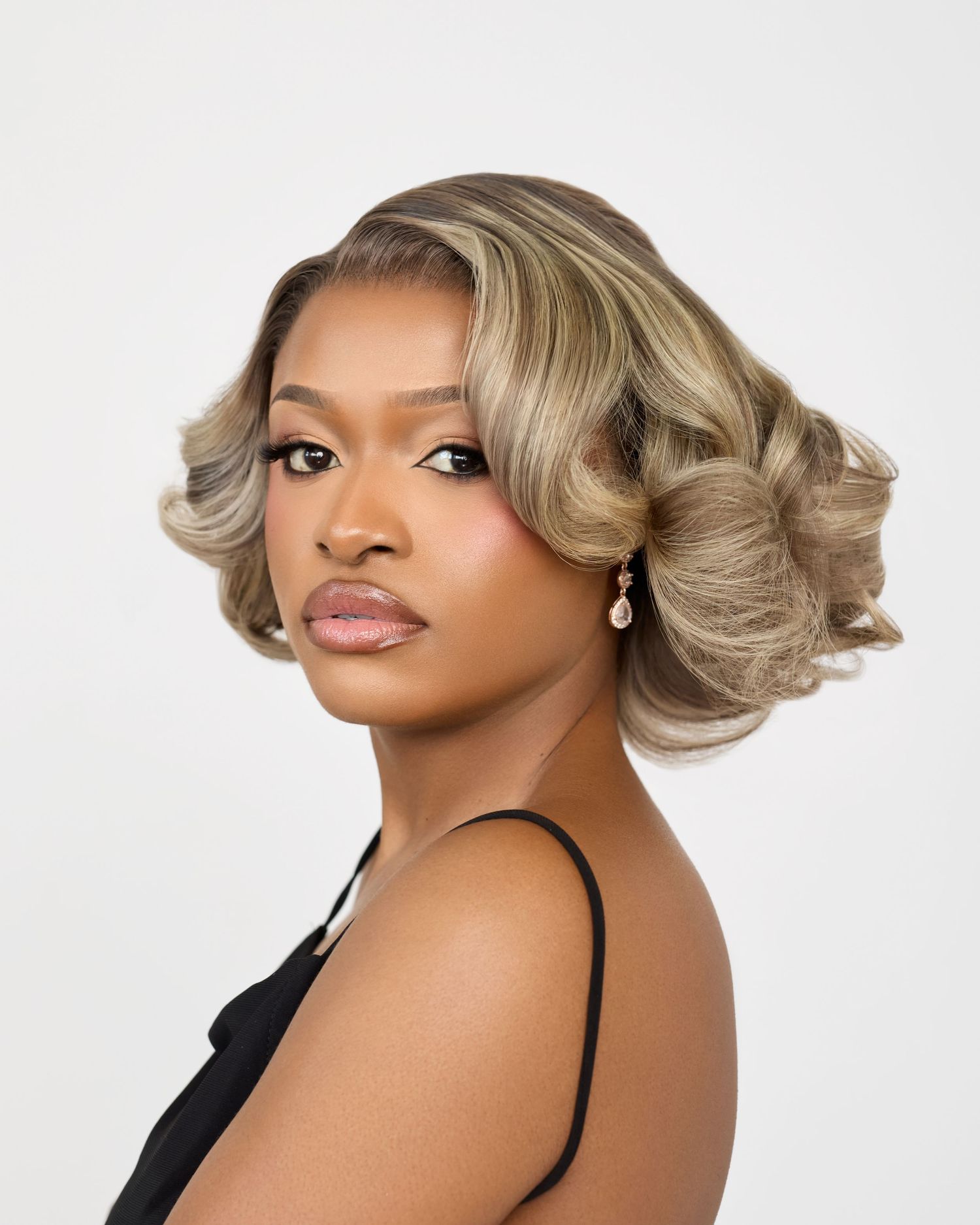 A young woman with elegant blonde highlighted waves, wearing a black dress and pearl earrings, posing against a white background.